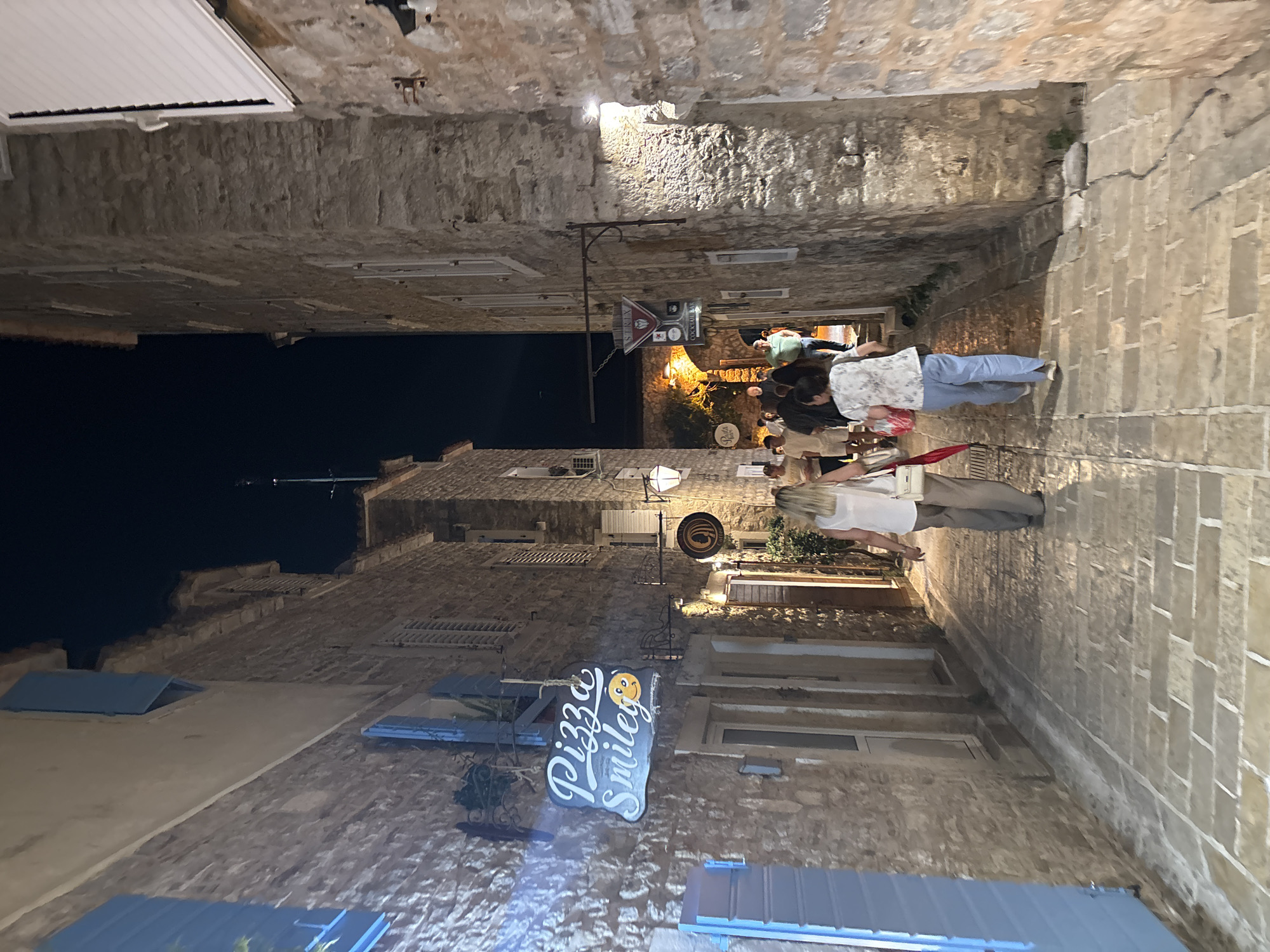 The image shows a narrow street at night in the old town of Budva, Montenegro. The street is paved with stone and lined with stone buildings on either side. A sign for "Pizza Smile" is visible. Several people are walking down the street. A restaurant or bar is visible in the background, illuminated by yellow lights. The sky above is dark.