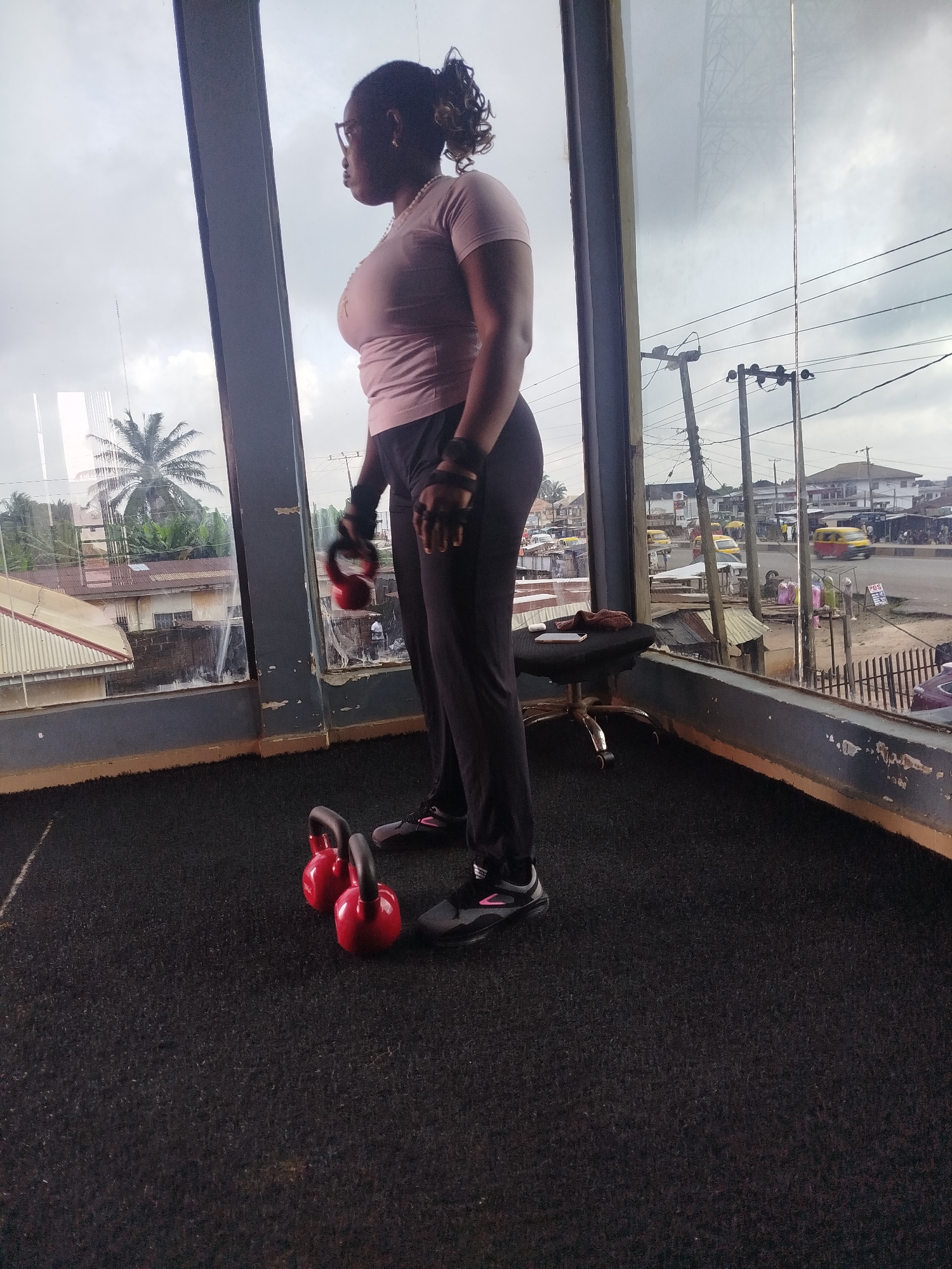 A woman is standing on a black mat in a gym, looking out of a large window at the city of Uselu, Nigeria. She is wearing a pink t-shirt, black pants, and black athletic shoes with pink accents. She is also wearing workout gloves. In front of her, on the mat, are three red kettlebells. The window shows a street scene with cars, buildings, and trees. The sky is overcast.