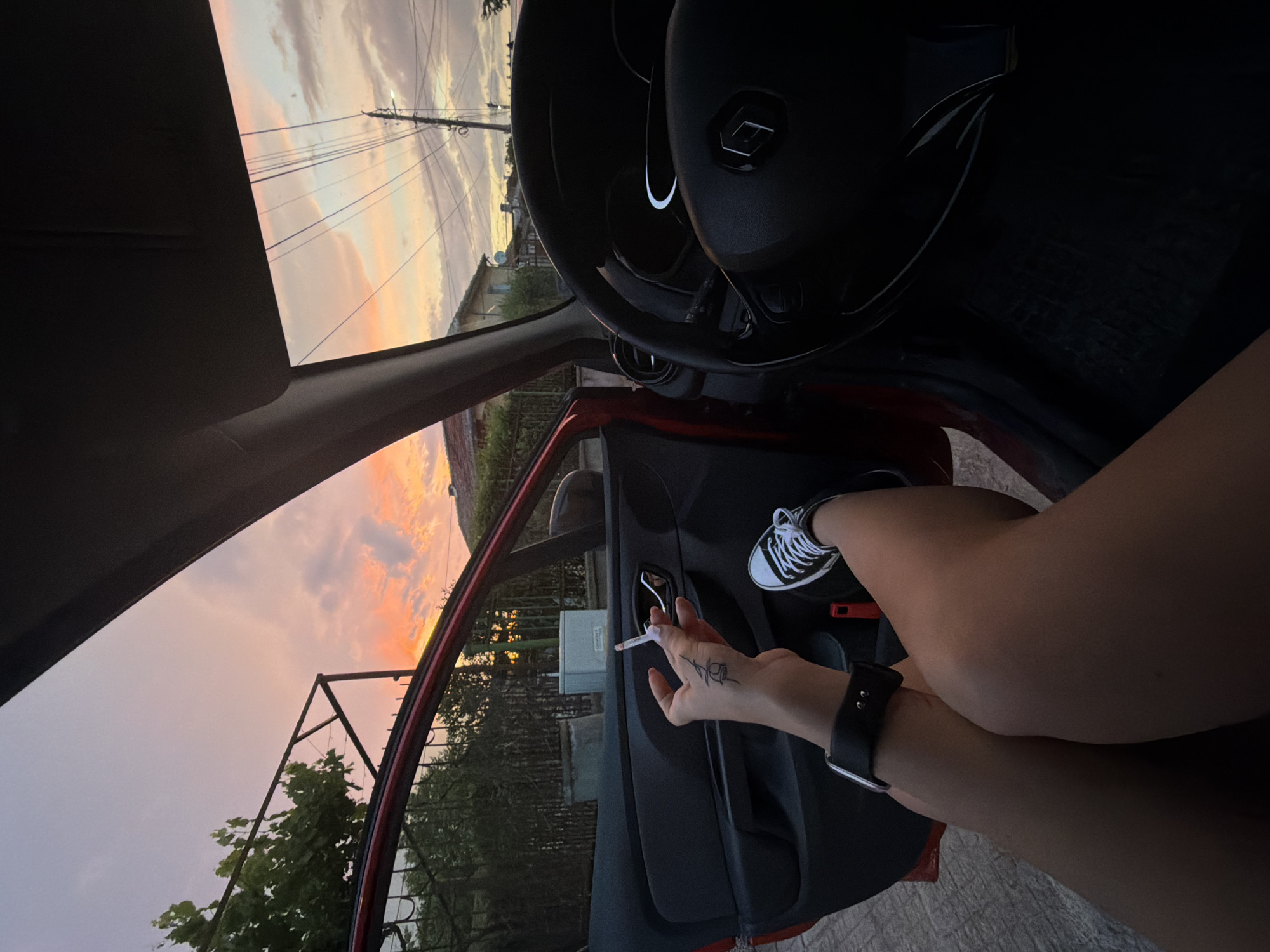 An exterior shot taken from inside a car shows a person's legs and hand holding a cigarette as the car door is open. The person is wearing sneakers and a wristwatch. The background features a sunset with clouds, houses, and power lines.