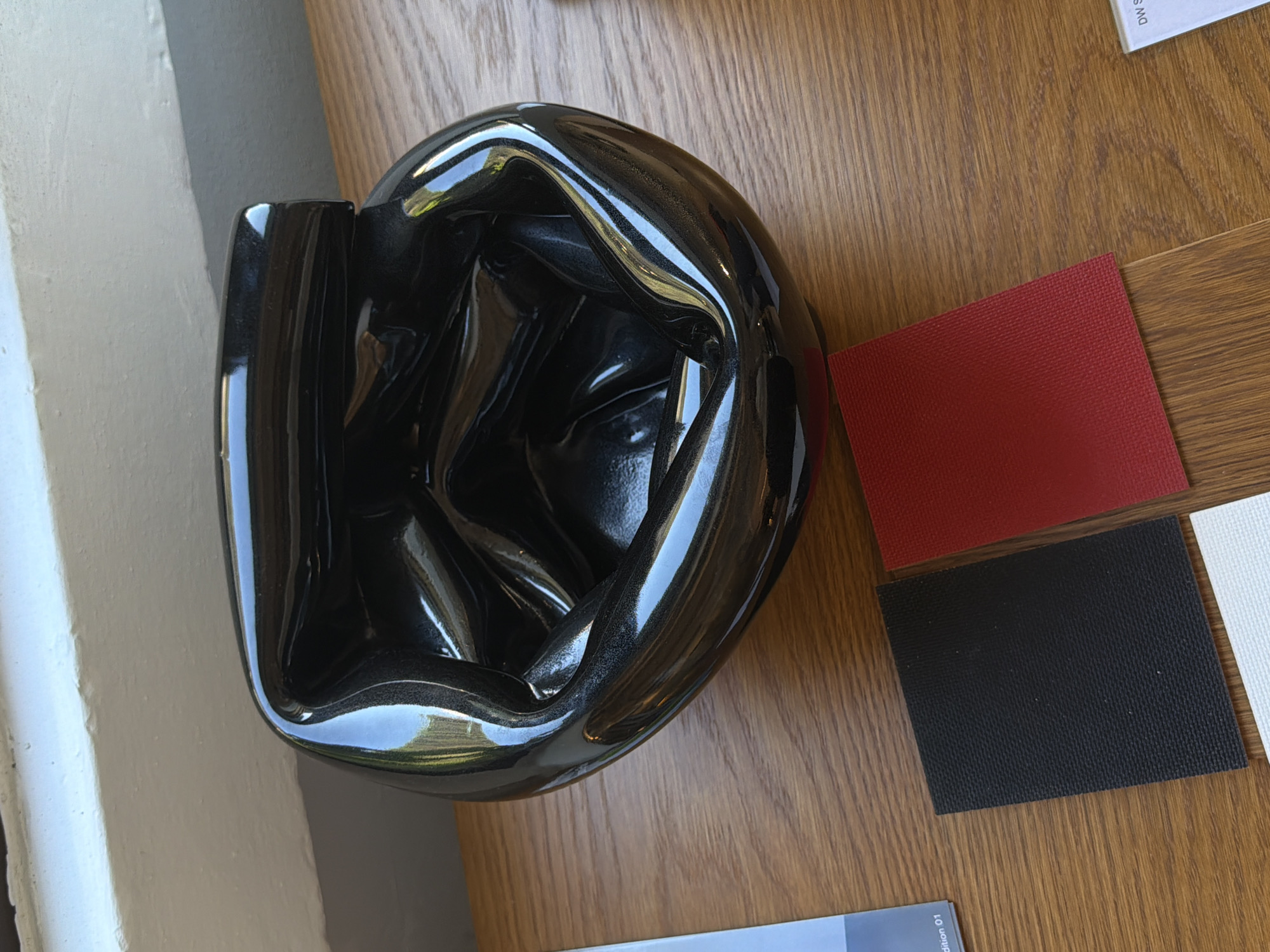An overhead shot captures a still life arrangement, likely within an interior design studio or showroom in Milan, Italy. The primary subject is a highly polished, dark gray to black, irregularly shaped object, possibly a vase or a decorative bowl. Its surface is extremely reflective, distorting and mirroring the surrounding environment, including a bright, light-colored wall or pillar to the left, and hints of green and blue, possibly from a window or outdoor view, are visible in the reflections. The object's form is fluid and sculptural, with undulating curves.
The object rests on a warm, medium-brown wooden surface with a distinct grain pattern, suggesting a table or desk. To the right of the object, three rectangular fabric swatches are arranged on the wooden surface. From top to bottom, these are a textured red swatch, a textured black swatch, and a partially visible textured white or off-white swatch.
In the background, at the top right, a piece of paper or card is partially visible with the text "DW S". Another piece of paper or card is seen at the bottom right, displaying "dition 01". The bright, even lighting suggests it is daytime. No people are visible, and the scene implies a professional setting for showcasing materials and design elements.