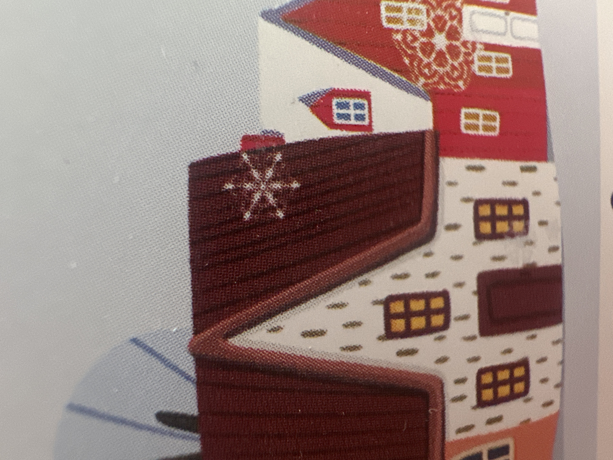 The image displays a close-up of a printed fabric or paper depicting a stylized cluster of buildings. The architecture is whimsical, with red roofs, white and red walls, and dark brown accents. Several windows are visible, depicted as small, square or rectangular shapes with yellow panes. One prominent element is a dark red wall with a white snowflake-like symbol. The buildings are stacked and angled, creating a sense of depth and complexity. The background is a plain, light gray texture, possibly representing sky or a neutral surface. The overall aesthetic is illustrative and decorative. There is no visible text in the image. The context of Minsk, Belarus, does not appear to be directly represented by any discernible landmarks or cultural cues within the image itself.