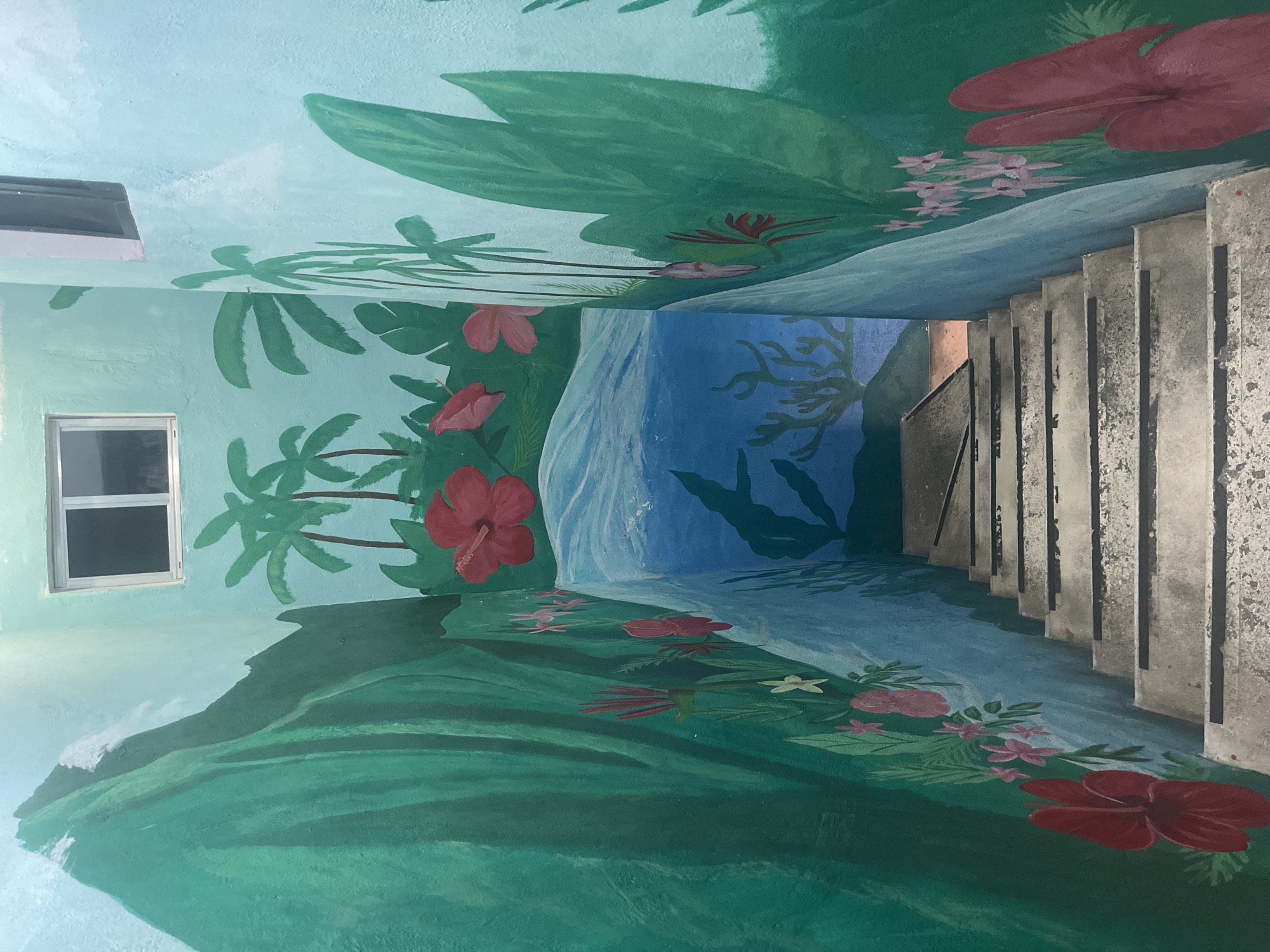 This image depicts a staircase decorated with a mural. The mural features a tropical scene with palm trees, hibiscus flowers, and lush green foliage painted on the walls and ceiling of the stairwell. A waterfall or a body of water is also part of the painting. The staircase itself is concrete and leads downward. The location is in the city of Playa del Carmen, Mexico.