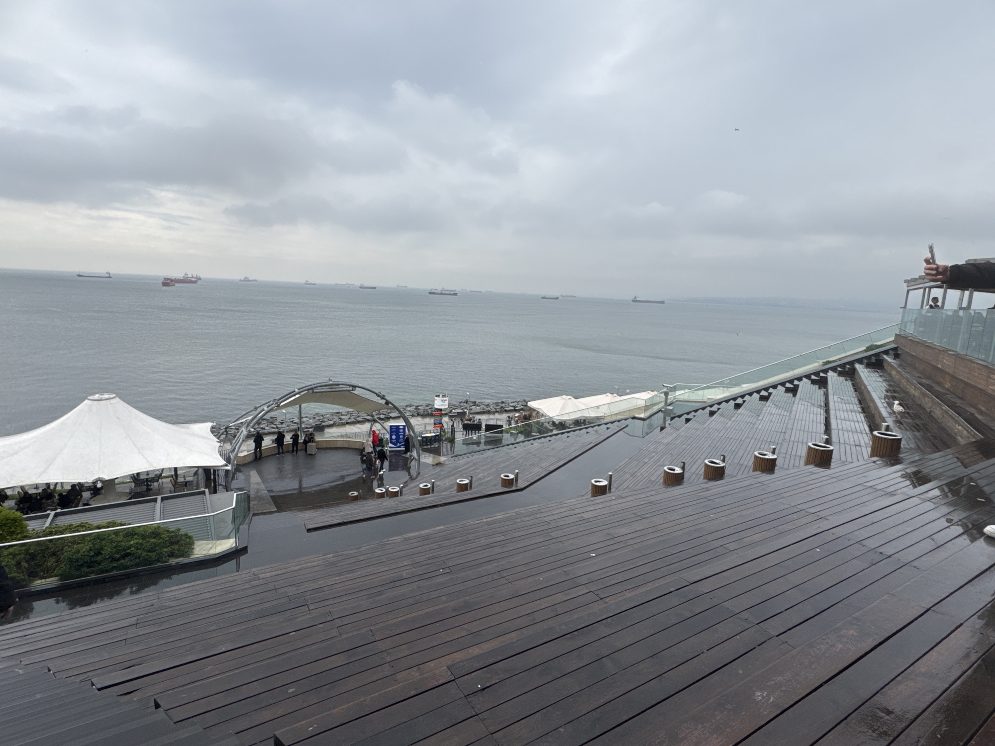 The image depicts a waterside location on a cloudy day in Bakırköy, Turkey. The foreground consists of a wide, stepped wooden deck that appears to be wet from rain. On the deck, there are cylindrical structures with metal tops spaced along its length. To the left, a white tent-like structure covers an area where people are seated at tables. Beyond this, a walkway leads to an observation area or platform with a curved, metal canopy.
In the background, numerous ships are visible on the water. The sky is overcast and gray, contributing to the somber atmosphere of the scene. The image suggests a public area designed for leisure and observation of the sea.