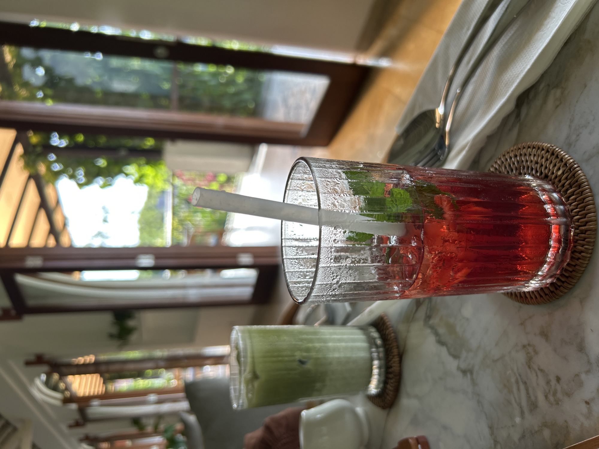 The image depicts two beverages on a marble table, likely a countertop. The beverage in the foreground is a red-colored liquid, possibly a juice or cocktail, served in a clear, textured glass with a white straw and some green herbs visible inside. The glass is placed on a small, round woven coaster. Behind it is a green-colored beverage, possibly a smoothie or iced matcha, also in a clear glass on a similar coaster. The lighting suggests an indoor setting with natural light, possibly near a window. Silverware wrapped in a white cloth napkin is visible next to the red drink. The background shows an out-of-focus view of what appears to be foliage and an open doorway or window, suggesting an open-air or semi-outdoor establishment. The location, Kuta Selatan, Indonesia, is provided but not directly visible in the image.