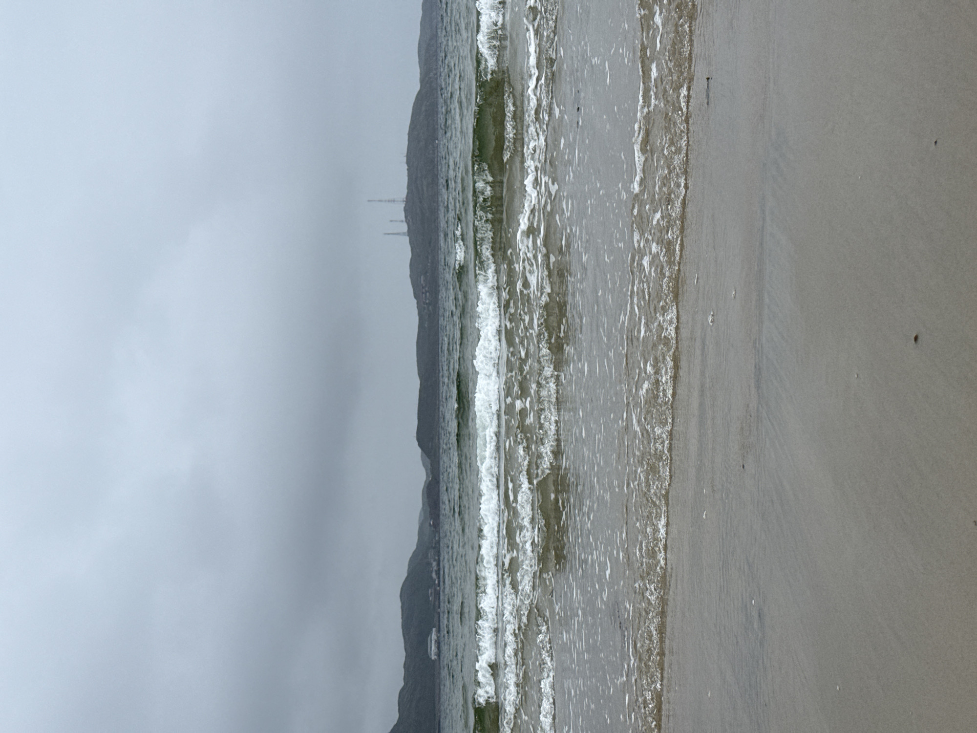 The image depicts a cloudy, overcast day at a beach in Teriberka, Russia. The foreground consists of wet sand with small shell fragments scattered on it. The middle ground is dominated by the ocean, with several rows of white-capped waves rolling towards the shore. In the background, a range of dark, rounded hills with some sparse vegetation is visible. On top of the hills, three tall transmission towers stand out against the muted sky, and some small buildings are clustered at the base of the hills. The overall atmosphere is somber and windswept, typical of a northern coastal environment. There are no people or animals visible in the frame.