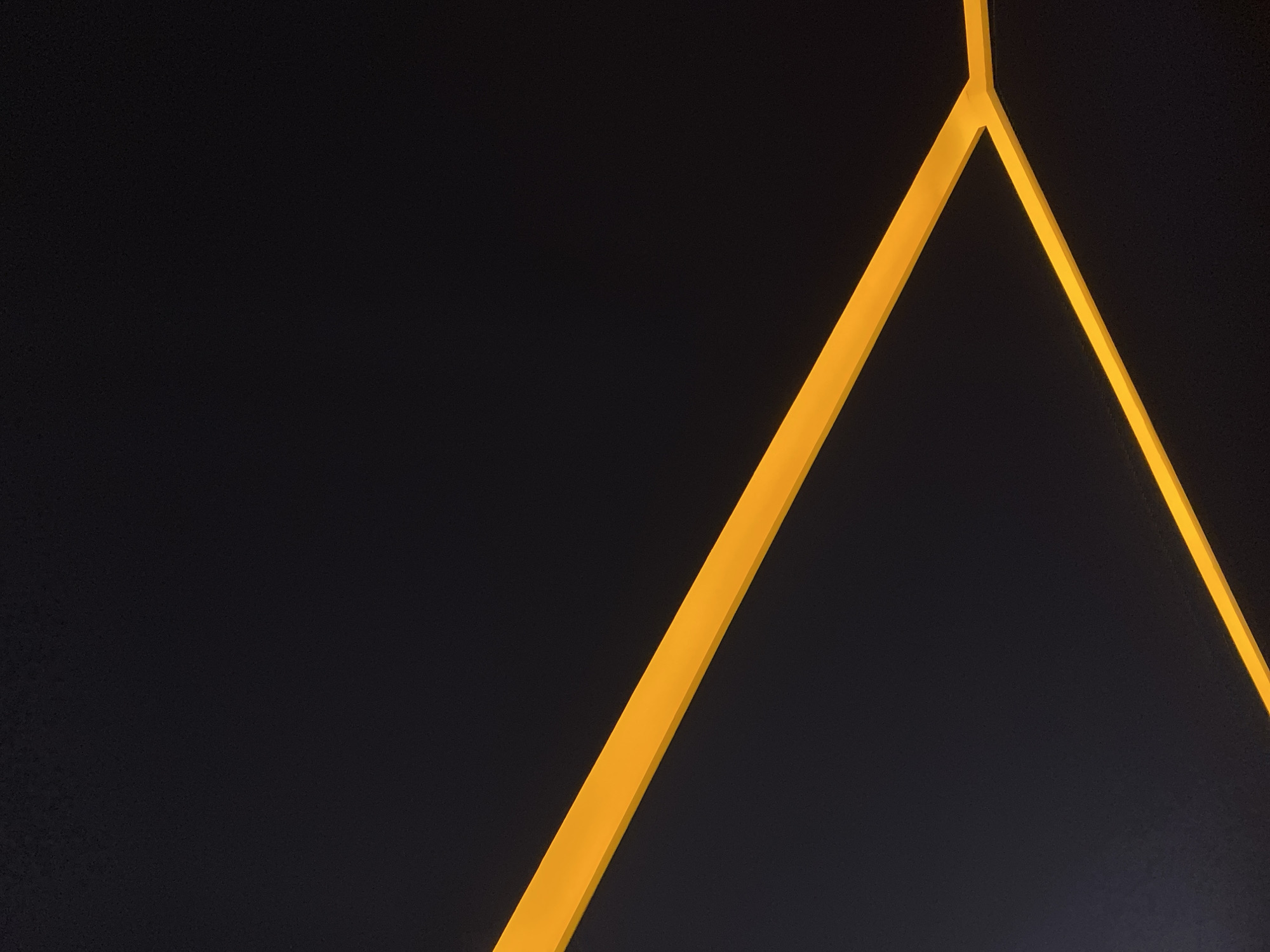 This minimalist and abstract image features bright, luminous yellow geometric lines set against a stark black background. The prominent lines converge towards the top-right portion of the frame, forming a distinct 'Y' shape. One thick, illuminated line runs diagonally from the lower-left corner upwards and to the right, meeting another narrower diagonal line extending from the upper-right towards the center. A short segment of the yellow line extends further upwards from this junction, exiting the top edge of the image.
The uniform, dark background provides a strong contrast, making the brightly colored lines appear to glow. The lines are straight and have crisp edges, suggesting a deliberate design, possibly an architectural element, a modern lighting fixture, or an abstract art installation. The overall impression is one of modernism and precision, with the focused illumination implying a dark environment, possibly indoors or at night. There are no identifiable objects, people, text, or specific location cues beyond the abstract geometric forms.
