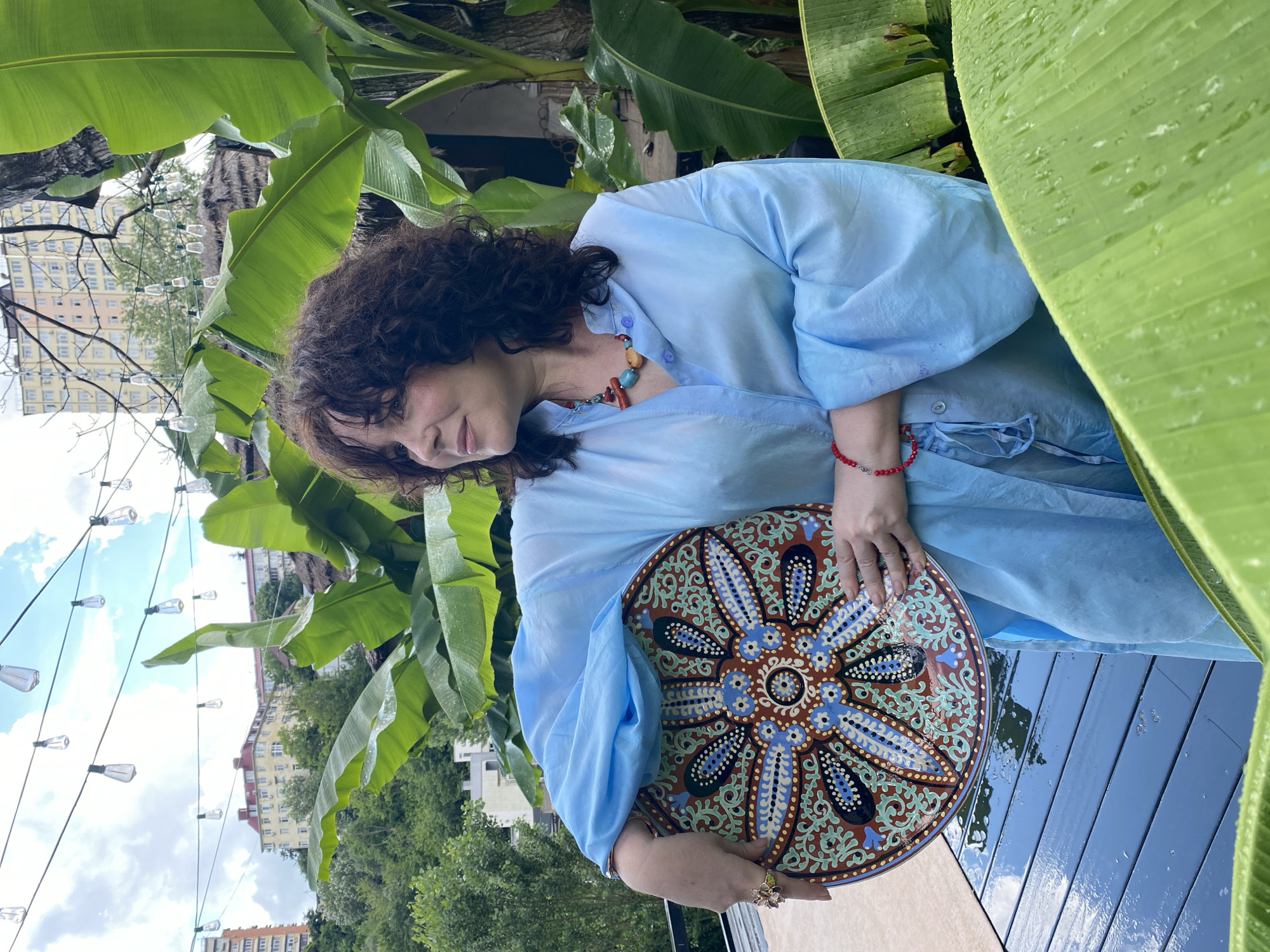 A woman with dark, curly hair is captured in a close-up shot, holding a decorative plate with intricate patterns. She is wearing a light blue, loose-fitting blouse and a colorful necklace. A red bracelet is visible on her left wrist, and she has a ring on her right hand. The woman's head is tilted down, and she appears to be looking at the plate, with a gentle, contemplative expression.
The setting suggests an outdoor environment, with large, lush green banana leaves dominating the foreground and framing the subject. Behind the woman, a cityscape is visible, with buildings and trees under a cloudy sky. Strings of fairy lights are strung across the background, adding a decorative element to the scene. The overall impression is one of a relaxed, perhaps artistic, moment outdoors. The wet appearance of the banana leaves and the deck suggest recent rain or dew, hinting at humid weather. The presence of the cityscape in Sochi, Russia, provides geographical context. The time of day is likely daytime, given the ambient light, but the cloudy conditions make it difficult to pinpoint precisely.
