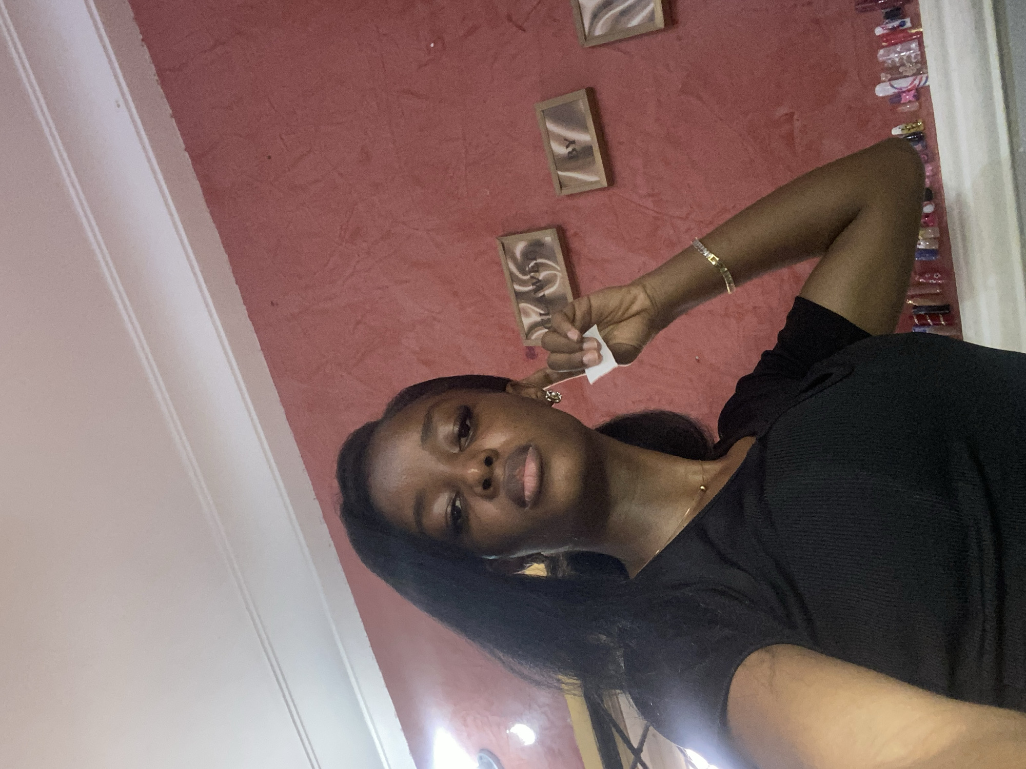In a selfie taken indoors, a young woman with dark skin and black hair stands against a textured red wall. She is wearing a black V-neck top and a delicate gold necklace. Her left hand is raised, holding a small white object and touching her ear. A silver bracelet adorns her right wrist. The background features three framed art pieces with swirling patterns on the wall, and a shelf below them displaying an assortment of colorful objects, possibly nail art or cosmetics. The ceiling above has white molding. The context of Jos, Nigeria, is not ascertainable from the visual information presented in the image.