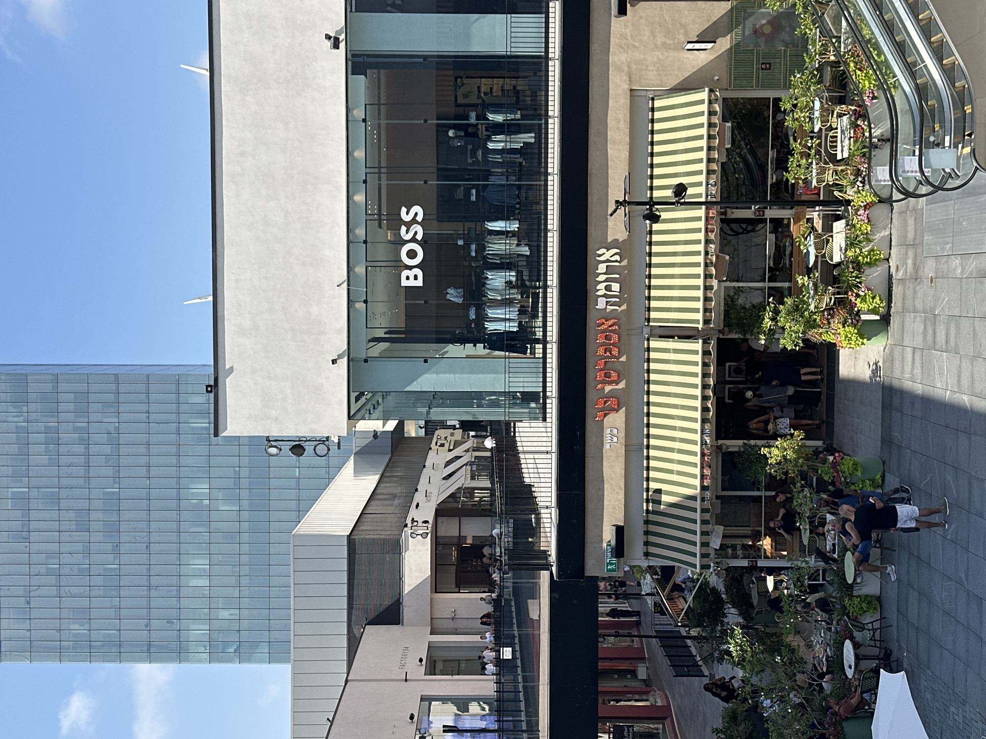The image shows a modern outdoor shopping area on a sunny day. In the foreground, a cafe with green and white striped awnings is visible, with tables and chairs set up outside amongst lush greenery. People are seated at some of the tables, and others are walking by. Escalators are on the right side of the frame. Above the cafe, a store with a large glass window displays clothing, with the word "BOSS" prominently displayed. In the background, a tall, glass-facade skyscraper dominates the skyline. Other buildings with modern architectural designs are also present. The scene appears to be in a bustling urban environment. Based on the Hebrew signage visible, the location is in Herzliya, Israel, likely in a contemporary shopping district such as the Arena Mall or a similar complex.