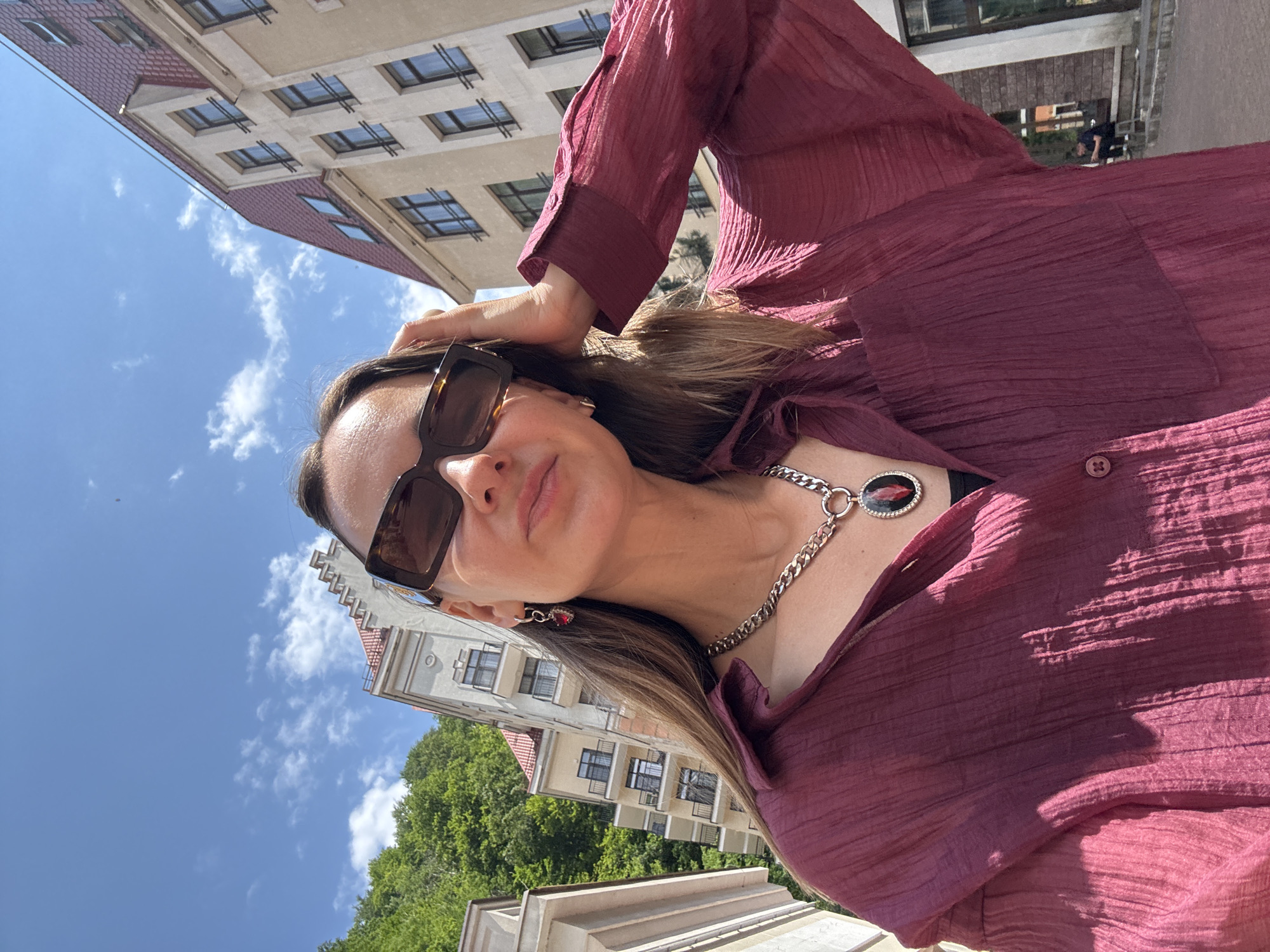 A woman is taking a selfie outdoors in Estosadok, Russia. She is wearing a burgundy collared shirt, sunglasses, and a necklace with a black and red pendant. The background shows buildings and trees under a blue sky with clouds. The woman is touching her hair.