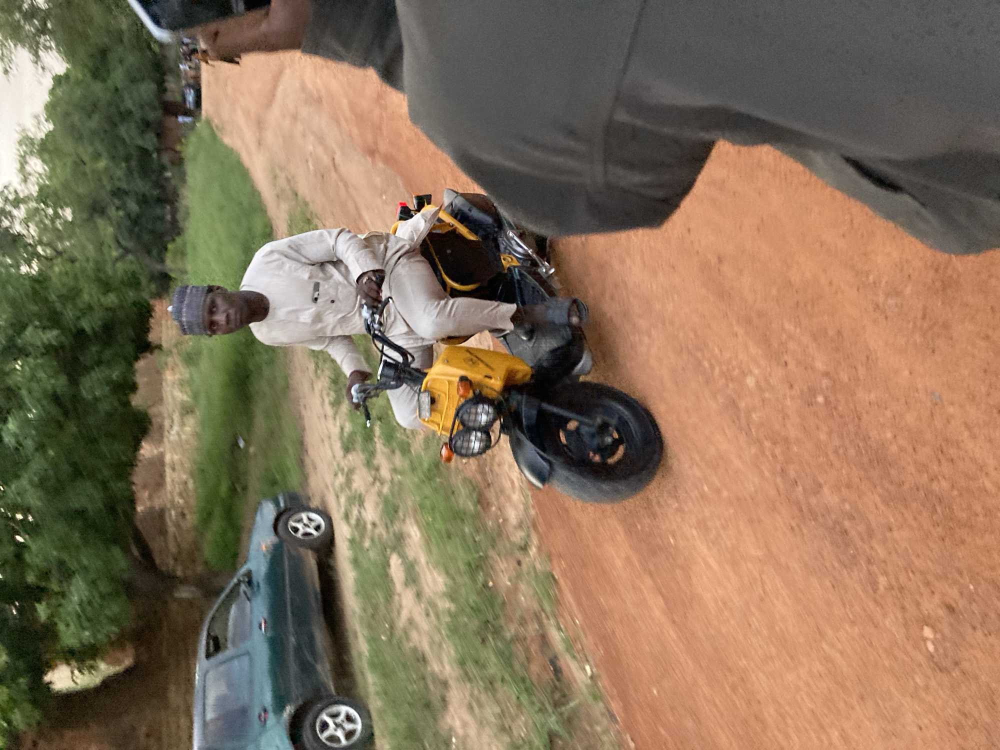 A man wearing a light-colored traditional outfit and a patterned cap is riding a yellow scooter on a dirt road. He is looking towards the camera. In the background, there is a dark green car parked on the side of the road, and lush green trees and foliage. The setting appears to be outdoors, with a rural or semi-rural feel. The overall lighting suggests it is late afternoon or early evening. The location is identified as Jos, Nigeria.
