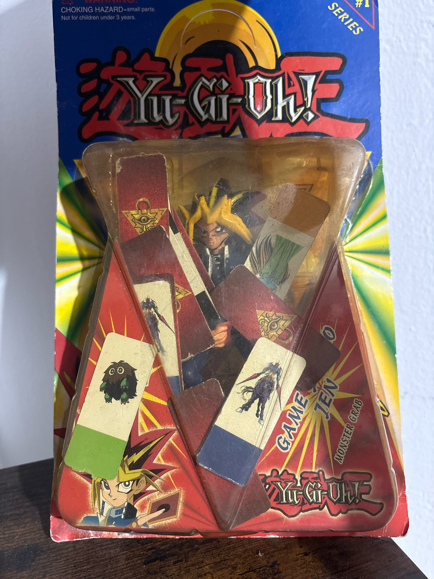 This image shows a Yu-Gi-Oh! toy package, likely from the early 2000s, featuring a figure of Yugi Muto and various collectible cards inside. The packaging is a clear plastic blister on a blue cardboard backing, which displays the Yu-Gi-Oh! logo and graphics. Inside the blister, several cards with illustrations of monsters and characters are visible, along with what appears to be a small figurine. The product is marked with "SERIES #1" and "CHOKING HAZARD—small parts. Not for children under 3 years." The image is taken from a slightly elevated angle, showing the toy package resting on a dark wooden surface. The background is a plain white wall with a hint of a bright yellow and green sunburst pattern visible behind the packaging. There is no information in the image to suggest it is located in Cuernavaca, Mexico.