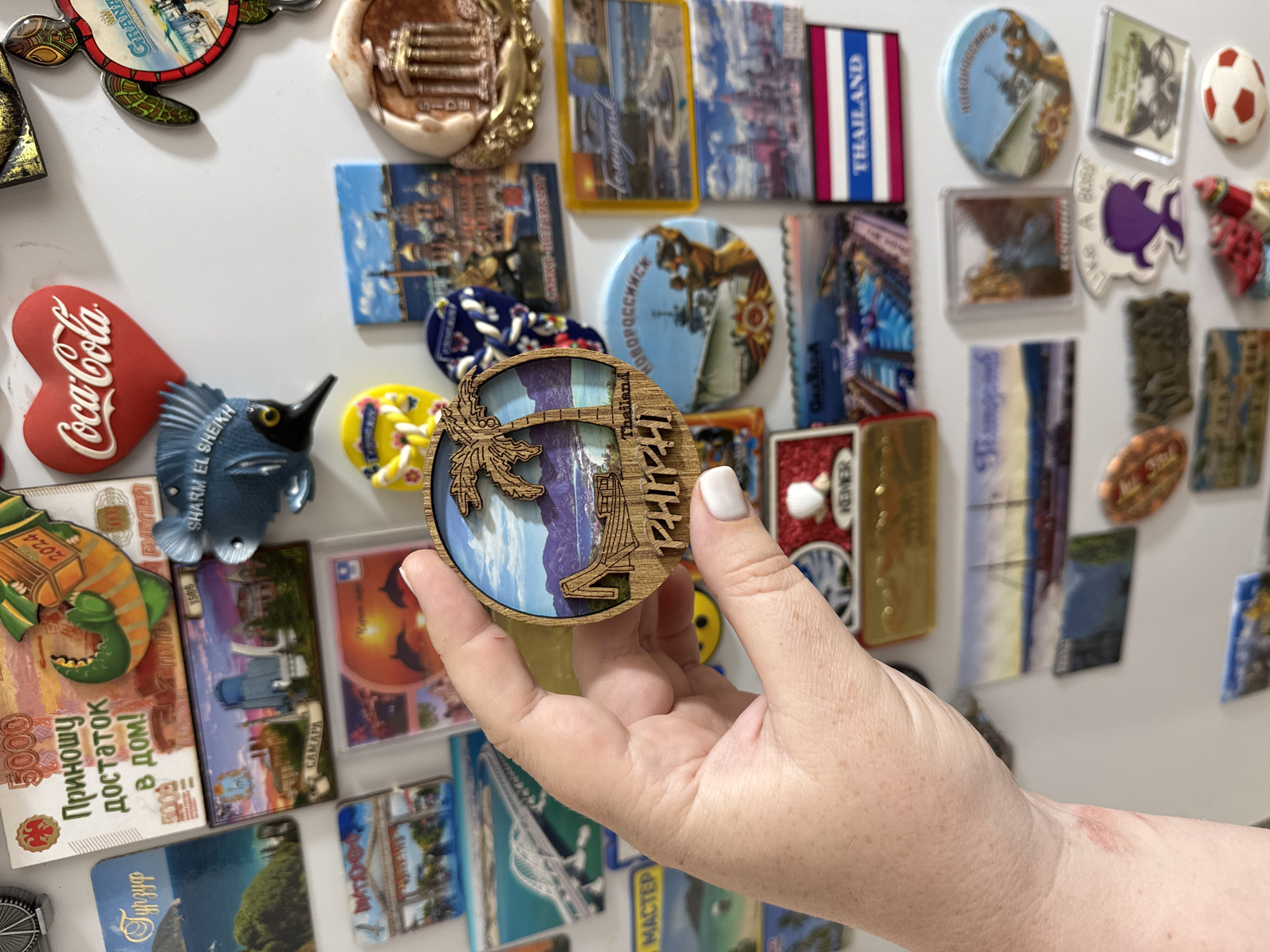 The image displays a collection of refrigerator magnets. The foreground features a person's hand holding a round, wooden magnet with a depiction of a palm tree and tropical scenery, along with the text "Thailand" and "PHIPHI." The background is a white surface, likely a refrigerator, covered with numerous other magnets. These magnets vary in shape, size, and design, featuring landmarks, text, and colorful illustrations. Some of the magnets are recognizable as representations of various locations. One magnet has text in Russian that appears to say "Приношу достаток в дом!" which translates to "I bring prosperity to the home!". Another magnet features the Coca-Cola logo. There are also magnets depicting buildings, bridges, and scenic landscapes. The image does not contain any information about the city of Mikhaylovsk, Russia.