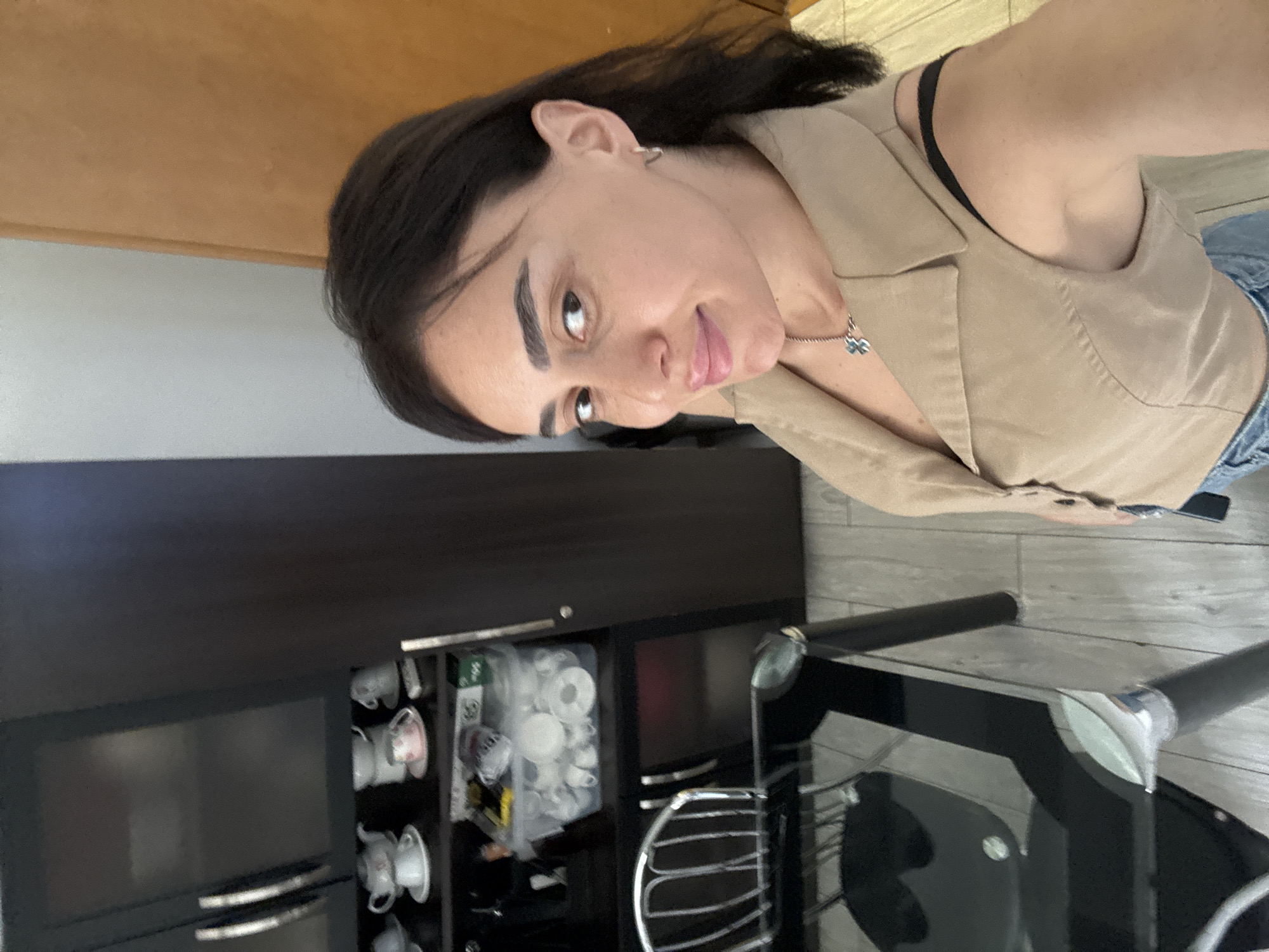 In a domestic interior, a woman with dark hair and brown eyes poses for a selfie, looking over her shoulder at the camera. She is wearing a beige sleeveless vest over a black garment and blue jeans. She has a light-colored necklace with a blue charm. Behind her, a dark wood cabinet is filled with various teacups and saucers. To her left, a modern glass-topped dining table with metallic legs is visible, accompanied by a metal-framed chair. The environment appears to be a home setting. The image appears to have been taken in Tbilisi, Georgia.