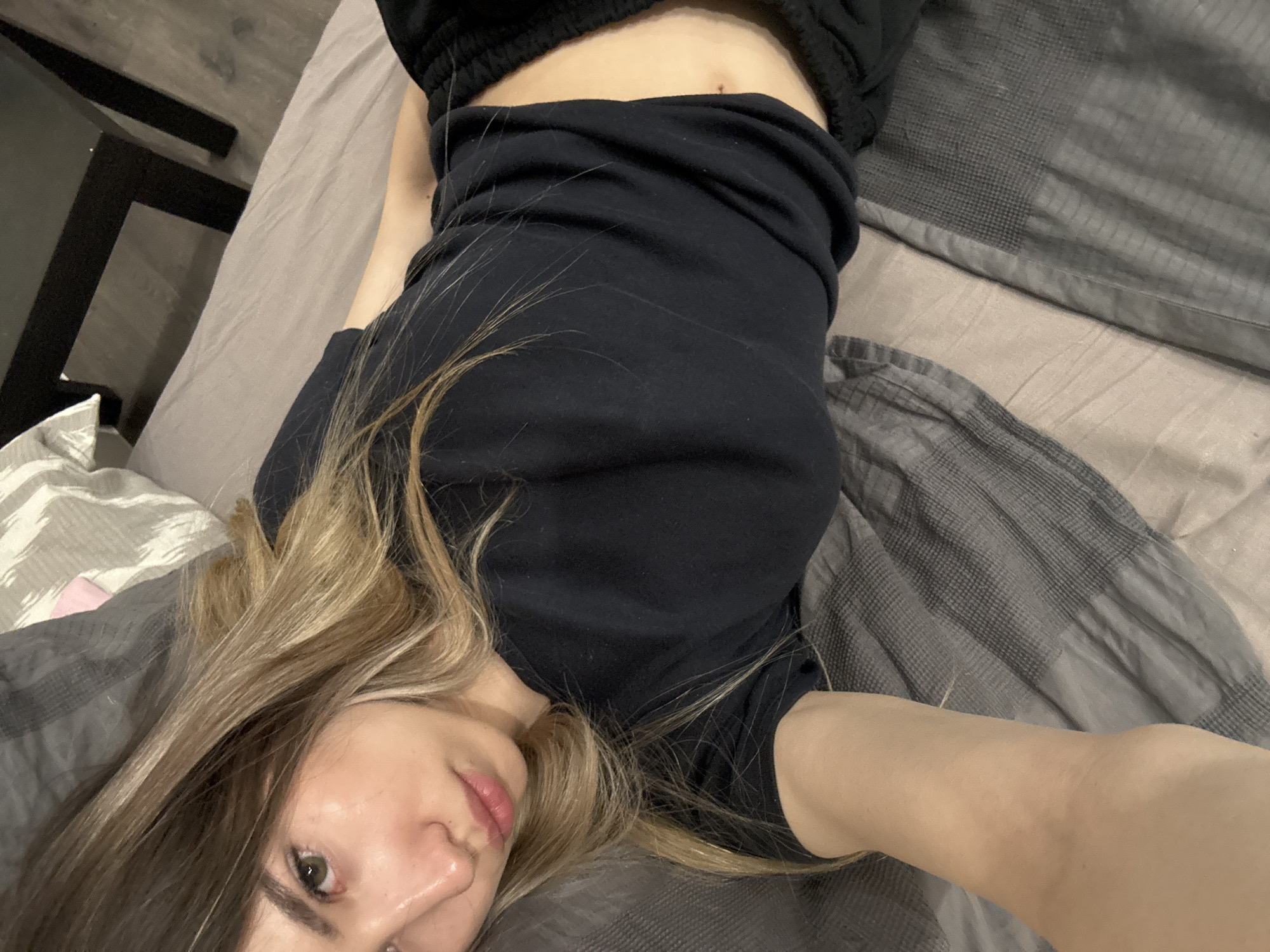 A woman lies on a bed covered with gray bedding. She wears a black t-shirt and black pants. Her blonde hair is partially spread on the pillow. The image appears to be a selfie taken from a low angle. The location is not identifiable as Chaikovsky, Russia.
