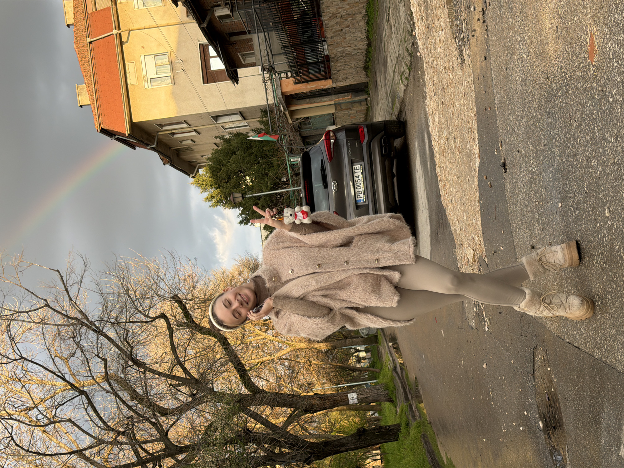 A young woman poses on a city street in Plovdiv, Bulgaria. She wears a light beige, fuzzy jacket, matching leggings, and boots. In her right hand, she holds a keychain with a small stuffed bear. She makes a peace sign with her left hand. A rainbow stretches across the sky in the upper left corner, ending near an old building with a red roof. Behind her, a dark grey car with a Bulgarian license plate (PB 0054 TE) is parked. A small Bulgarian flag is visible near the car. Bare tree branches frame the lower left portion of the image, while the wet asphalt street fills the foreground.