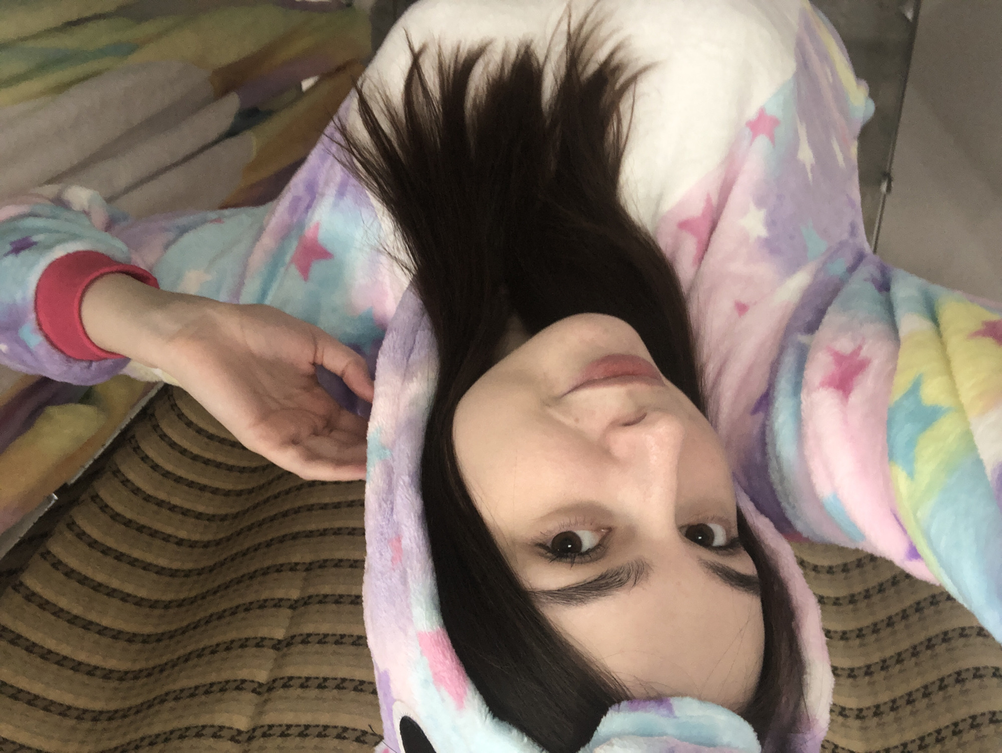 A young woman lies on a bed in a brightly lit room. She is wearing a light blue and pink unicorn onesie with a hood. Her long, dark hair spreads out on the white fabric around her head. The bed has a brown striped pattern. The location cannot be determined from the image.