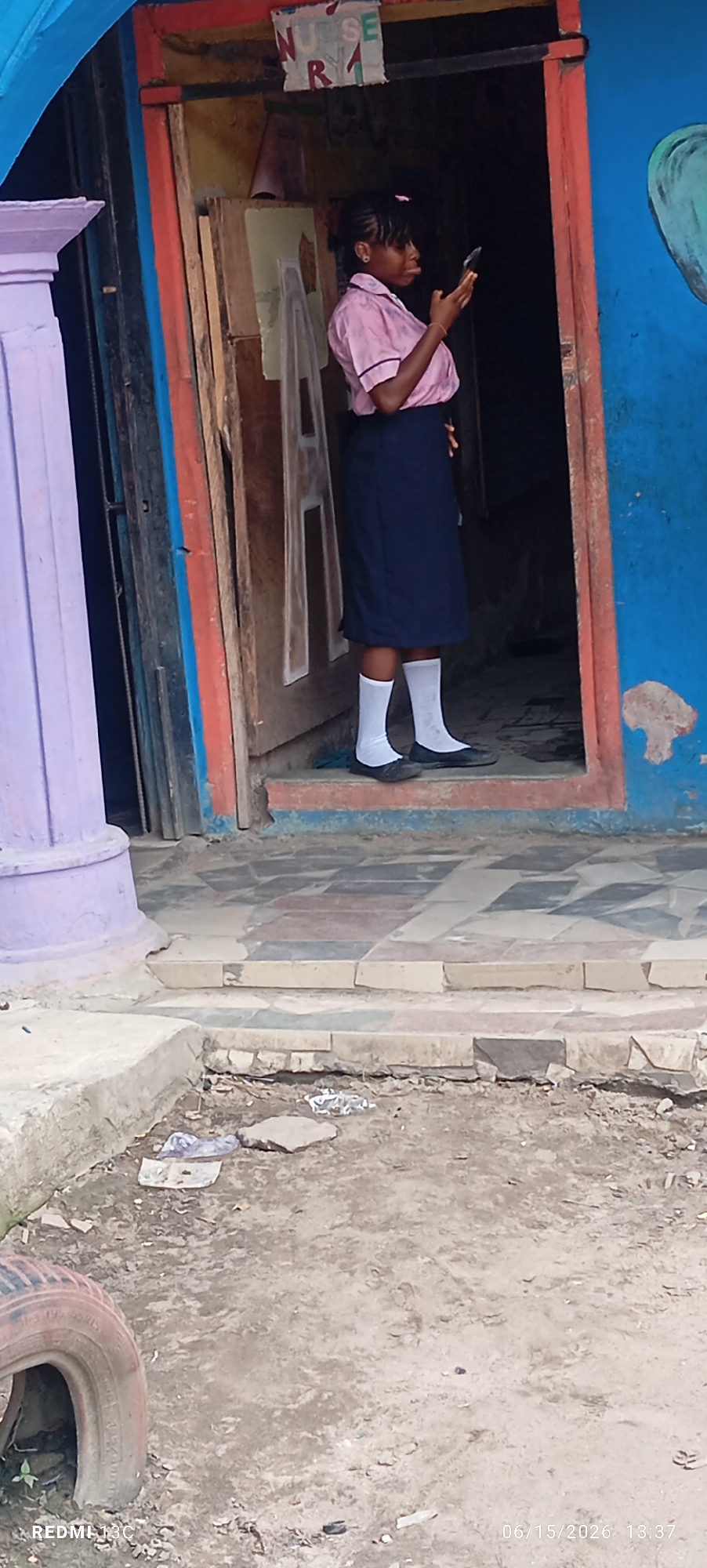 In Lagos, Nigeria, a young woman stands in the doorway of a brightly painted building. She is wearing a pink polo shirt with a dark navy skirt and white socks with black shoes, suggesting she may be in uniform. Her hair is braided and pulled back. She is looking down at her mobile phone, holding it in her right hand. The building's entrance is framed by a blue wall on the right and a red-painted frame. A large, light purple pillar stands to the left of the entrance. Inside the doorway, there are wooden panels and what appears to be a sign with the letters "NURSE" and "R1" on it. The ground in front of the building is tiled with a pattern of squares in shades of beige, red, and gray. Further out, the ground is unpaved and appears to be dirt with some debris, including a discarded tire on the lower left. The overall scene depicts a street-level view of a commercial or residential building entrance in an urban Nigerian setting.