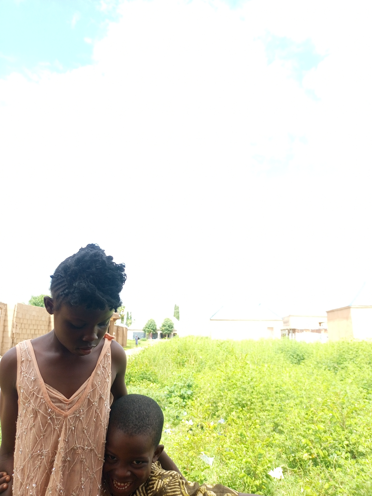 In Tempure, Nigeria, two young children are pictured outdoors against a backdrop of bright sky and lush green vegetation. The elder child, a girl, stands with her head bowed, wearing a sleeveless light peach dress adorned with intricate beadwork. Beside her, a younger boy is positioned in front of her, his face turned towards the camera. He has a broad, toothy smile and appears to be wearing a patterned brown and yellow shirt. Behind them, a few buildings with light-colored walls and roofs are visible, suggesting a semi-urban or rural setting. The overall scene is illuminated by bright daylight.