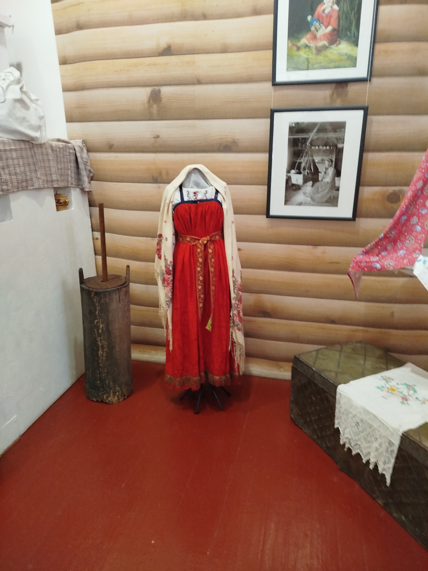 The image depicts an interior scene, likely within a museum or historical exhibit, featuring elements of traditional Russian culture. The walls are paneled with light-colored wood, creating a rustic ambiance. On the left, a white stove-like structure is partially visible, draped with a plaid cloth. Next to it stands a tall, cylindrical wooden object with a vertical handle, resembling a churn or a mortar.
Dominating the center of the frame is a mannequin dressed in a vibrant red traditional Russian dress, possibly a sarafan, cinched at the waist with a patterned belt. A light-colored shawl with floral embroidery is draped over the mannequin's shoulders and head.
In the background, two framed photographs hang on the wooden wall. The upper photograph appears to be a colored painting of a person in traditional attire, while the lower one is a black and white image of people in what looks like an interior setting.
To the right, a large, dark wooden chest rests on the red floor. A white embroidered cloth is laid over the chest. A section of a red and white patterned fabric, possibly a scarf or part of another garment, is draped on the wall to the right of the photographs. The floor is a uniform, dark red color.
The scene suggests a historical display related to Russian folk life. Without specific visual cues directly indicating the city of Novokuznetsk, Russia, its location cannot be definitively determined from the image alone.