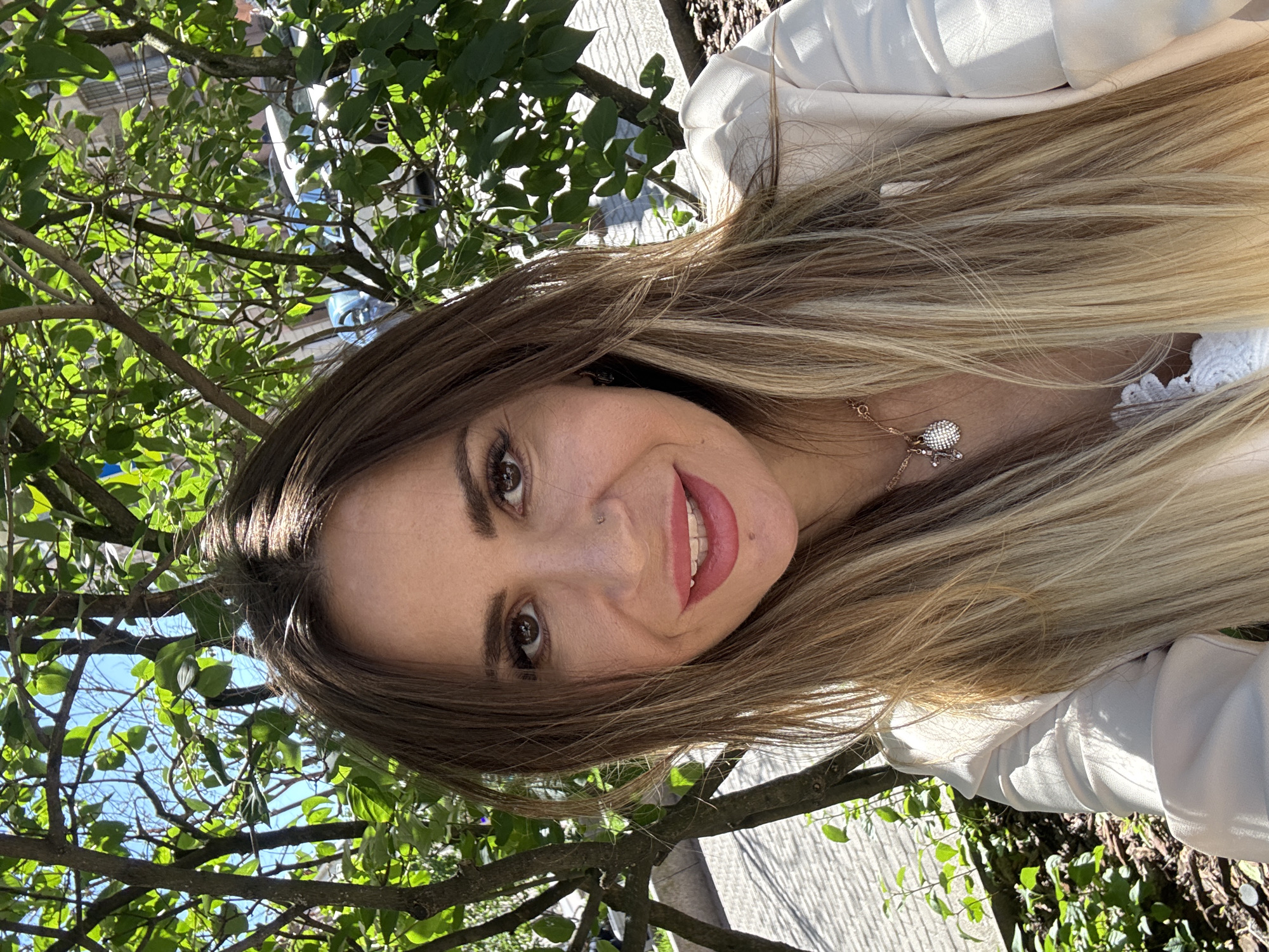 A blonde woman smiles for a selfie in an outdoor urban setting in Saint Petersburg, Russia. She has long, wavy blonde hair and is wearing a light-colored blazer over a white lace top. She has makeup on, including bright pink lipstick, and is wearing a necklace with two charms. She has a nose ring and a mole on her chin. The background shows green trees and branches, with a sidewalk or paved area and buildings visible through the foliage. The sun is shining, casting shadows from the trees onto the ground.