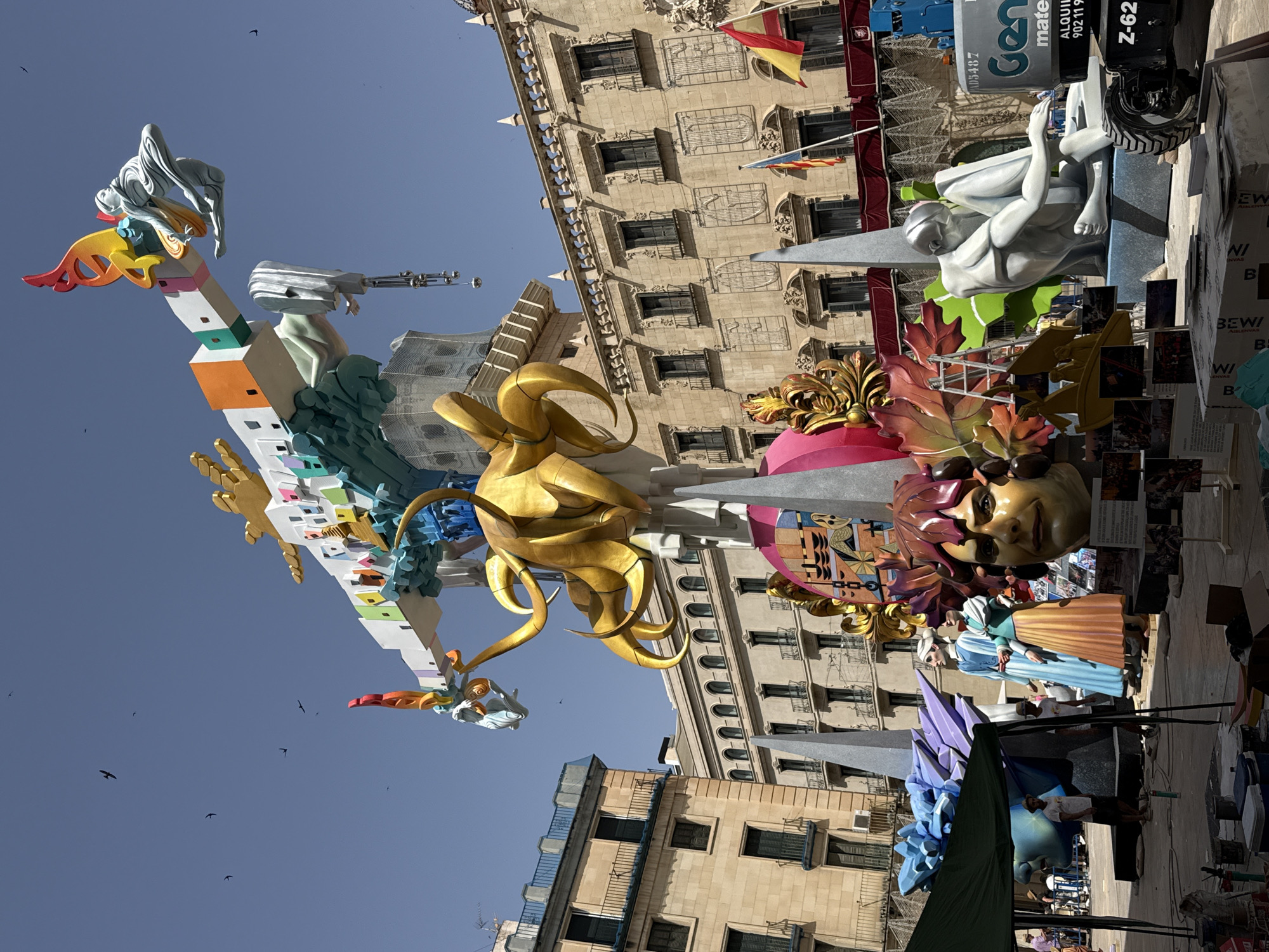 This is a vertical, eye-level, medium shot of a large, colorful, and complex effigy or sculpture set up outdoors in front of several old stone buildings. The sculpture is very tall and elaborate, featuring a mix of abstract shapes, figures, and architectural elements.
The subjects of the sculpture include a reclining white figure with smooth, sculpted lines, a large golden abstract form resembling tentacles or swirling fabric, and a giant face with dramatic makeup. There are also smaller figures, including what appear to be two women in traditional clothing. The sculpture is made of various materials, including what looks like painted wood or fiberglass, with bright, contrasting colors like gold, red, orange, yellow, blue, green, and white. The base of the sculpture is decorated with colorful blocks and a large, spiky blue structure.
The scene is set in a city, as indicated by the buildings in the background. The buildings are made of stone with arched windows and decorative architectural details. Flags are visible on some of the buildings, suggesting a public event or celebration. The sky is a clear, bright blue with a few small birds flying. The lighting suggests it is daytime, likely sunny, possibly mid-morning or afternoon.
The overall impression is one of a vibrant, artistic, and festive event. The sculpture appears to be part of a parade, festival, or exhibition, common in some Spanish-speaking cultures (like the Las Fallas festival in Valencia). Some visible text on a cylindrical object in the upper right corner includes "Gen", "mate", "ALQUIL" (likely short for alquiler, meaning rental), and a license plate "Z-62". The text "BEWI" is also visible on printed materials. The absence of specific landmarks makes the exact location unknown, but the architectural style and flags hint at a European setting.