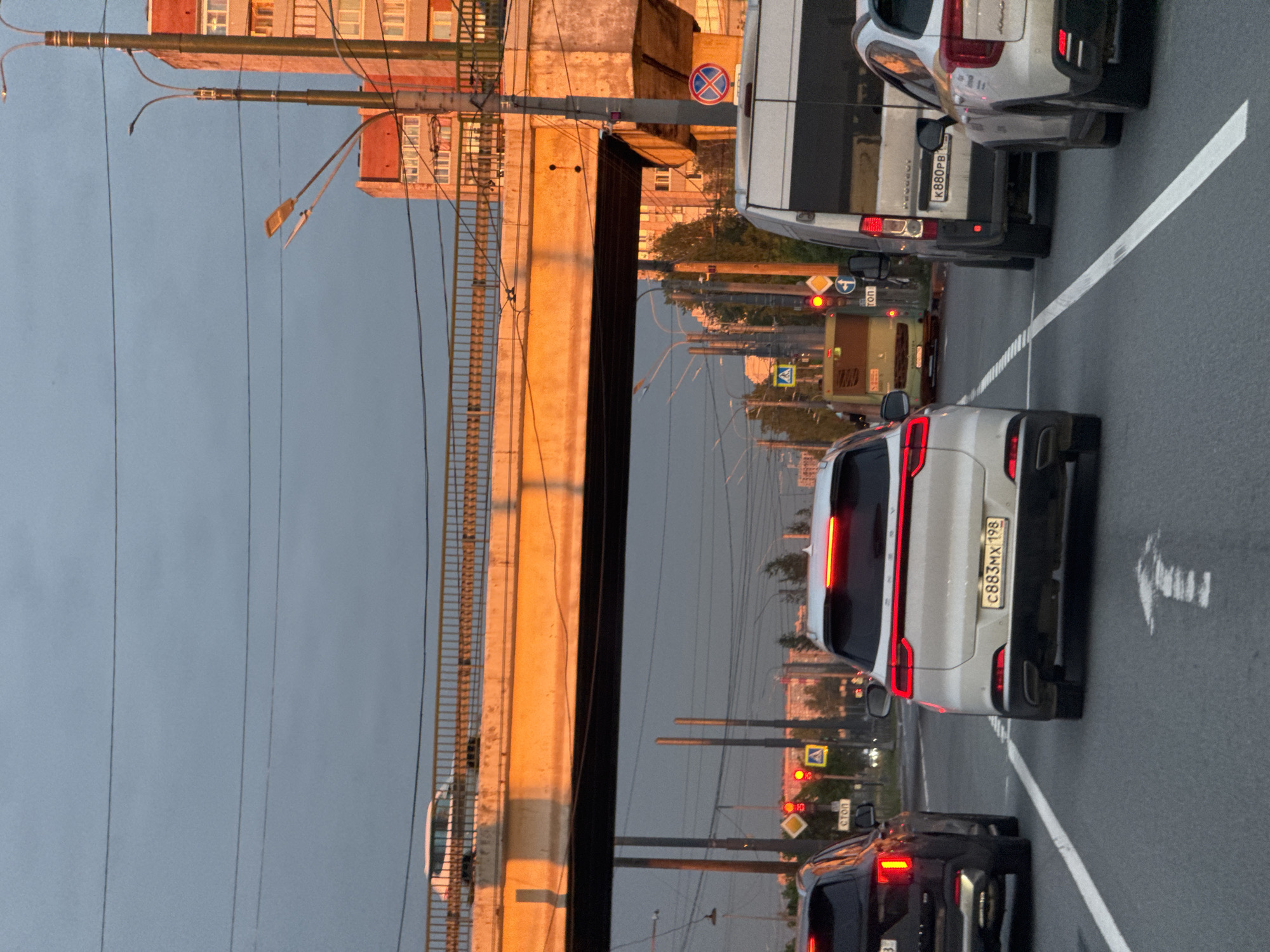 This image captures a busy road scene during the golden hour, likely late afternoon or early evening, characterized by warm, low-angle sunlight. Vehicles are stationary in traffic on a multi-lane road, indicating a halt at a traffic signal.
**Subjects:** The primary subjects are several vehicles in a traffic jam. In the foreground, a white SUV is visible, followed by a darker sedan. Behind them is a yellow public transport vehicle, likely a bus or trolleybus, and further back, a white van and another car. No people are distinctly visible.
**Scene and Setting:** The road features clear white lane markings, including a solid line separating the lanes and a white arrow painted on the asphalt, indicating a straight-ahead movement. To the left of the road, a substantial concrete structure, possibly a bridge or overpass support, is brightly illuminated by the golden light, creating a strong contrast with the cooler, shadowed areas. Above the road, a complex network of overhead power lines and lampposts is visible against a muted sky. In the background, partially obscured by the bridge and sparse foliage, are buildings with numerous windows, suggesting residential or commercial structures. Traffic lights show red, indicating vehicles are stopped.
**Activity:** The vehicles are at a standstill, suggesting a traffic jam or waiting at a red light. Brake lights are illuminated on several cars.
**Notable Details:** The warm, directional sunlight highlights the texture of the concrete structure and the buildings, creating a vivid contrast with the generally cooler tones of the sky and shadowed parts of the scene. The dense urban infrastructure, including multiple power lines and traffic signals, is prominent.
**Visible Text:**
* On the rear of the white SUV, the Russian license plate "C883MX 198" is visible.
* A white van further up the road displays the Russian license plate "K880PB 98".
* On the side of the yellow public transport vehicle, the Russian word "CTON" (meaning "STOP") is discernible.
* Traffic signs include a red circular sign with a diagonal cross (indicating "no stopping or standing"), a yellow triangular sign with a black pedestrian symbol (pedestrian crossing ahead), and a yellow diamond-shaped sign, often indicating a main road or priority.