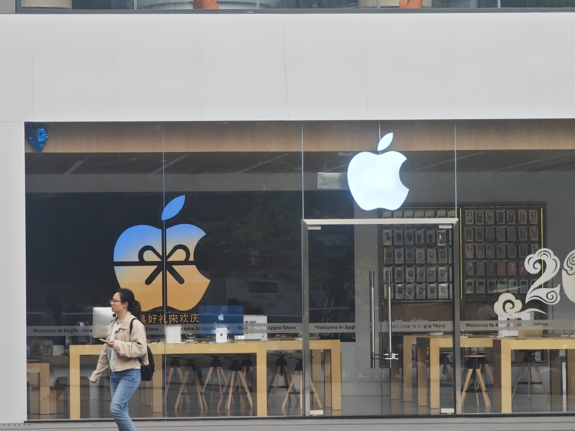 A daylight street-level view captures a modern Apple Store storefront in Yuzhong District, China. The store features a large glass facade, reflecting parts of the exterior environment. A woman in glasses, a light-colored jacket, blue jeans, and a dark backpack walks from left to right past the store, holding a white smartphone.
Inside the store, minimalist light wood tables with stools are set up, displaying Apple computers like iMacs. The interior lighting is bright. On the right side of the facade, a large, illuminated white Apple logo is prominently displayed. To the left, another large Apple logo is visible on a digital display or screen, designed with a blue top half, an orange bottom half, and a black ribbon creating a bow, giving the impression of a wrapped gift, suggesting a celebratory theme. A small blue leaf shape is also visible on the far left of the facade. Merchandise, likely phone cases, is displayed in neat rows on the back walls of the store.
Visible text on the glass includes "Welcome to Apple Store," repeated several times in English. Below the gift-themed Apple logo, Chinese characters read "美好礼来欢庆" (Měihǎo lǐ lái huānqìng), meaning "Good gifts come for celebration." To the far right, a large white decorative number "2" is partially visible, possibly part of a larger numerical display like "202x".