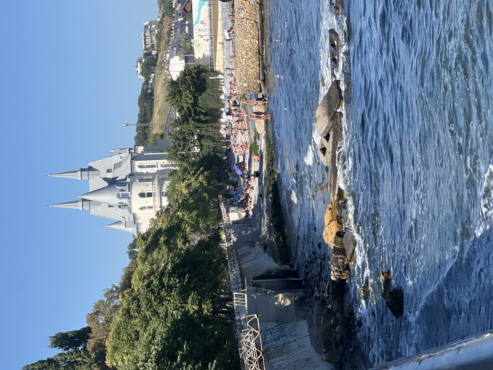 This is a photograph of the Odesa coastline in Ukraine. In the background, a fairytale-like white building with multiple spires sits on a hill, surrounded by lush green trees. Below the building, a lively beach scene unfolds with numerous people sunbathing, relaxing on towels, and swimming in the clear blue sea. A stone wall separates the beach from the water, with some people wading in the shallow areas. The foreground is dominated by the dark blue sea, with waves gently lapping against the shore and some weathered rocks and concrete structures emerging from the water. The sky is a clear, bright blue, indicating a sunny day. The overall impression is one of a popular tourist destination on a warm, summer day.