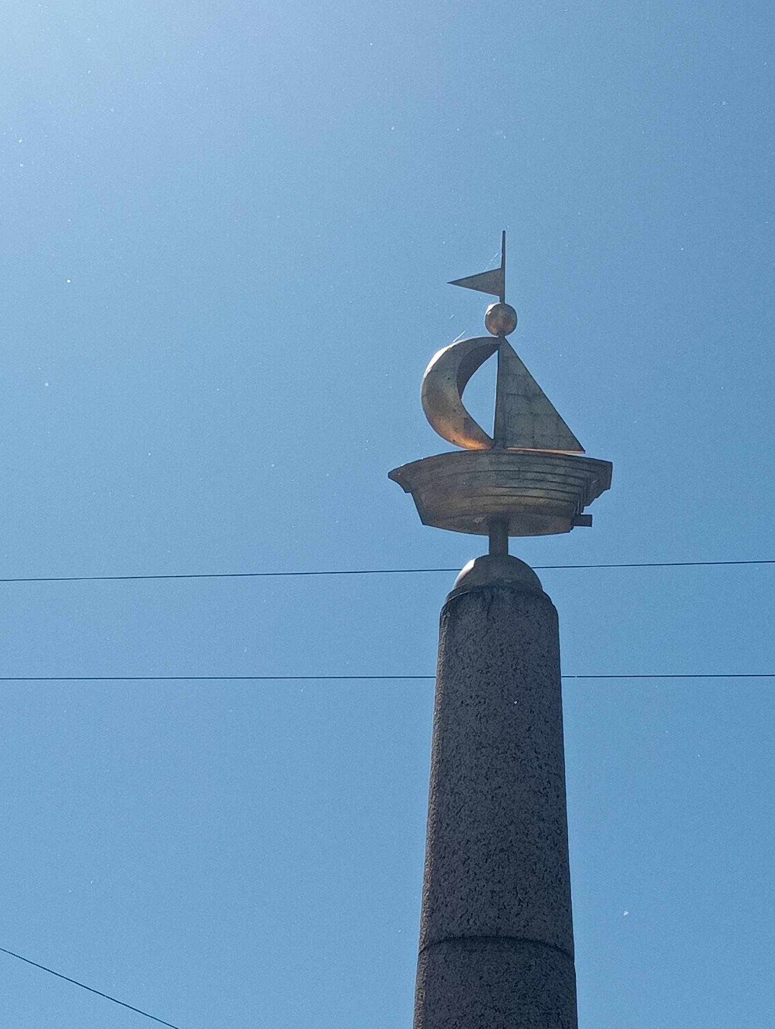 The image shows a golden sculpture of a sailing ship atop a granite obelisk. The sculpture features a ship's hull, a sail, and a mast with a flag. The obelisk is textured and has a band around its base. In the background, there is a clear blue sky with some faint lines that appear to be power lines. This is the V.Ya. Vasilevsky monument, located on Vasilievsky Island in Saint Petersburg, Russia, marking the entrance to the Neva River.