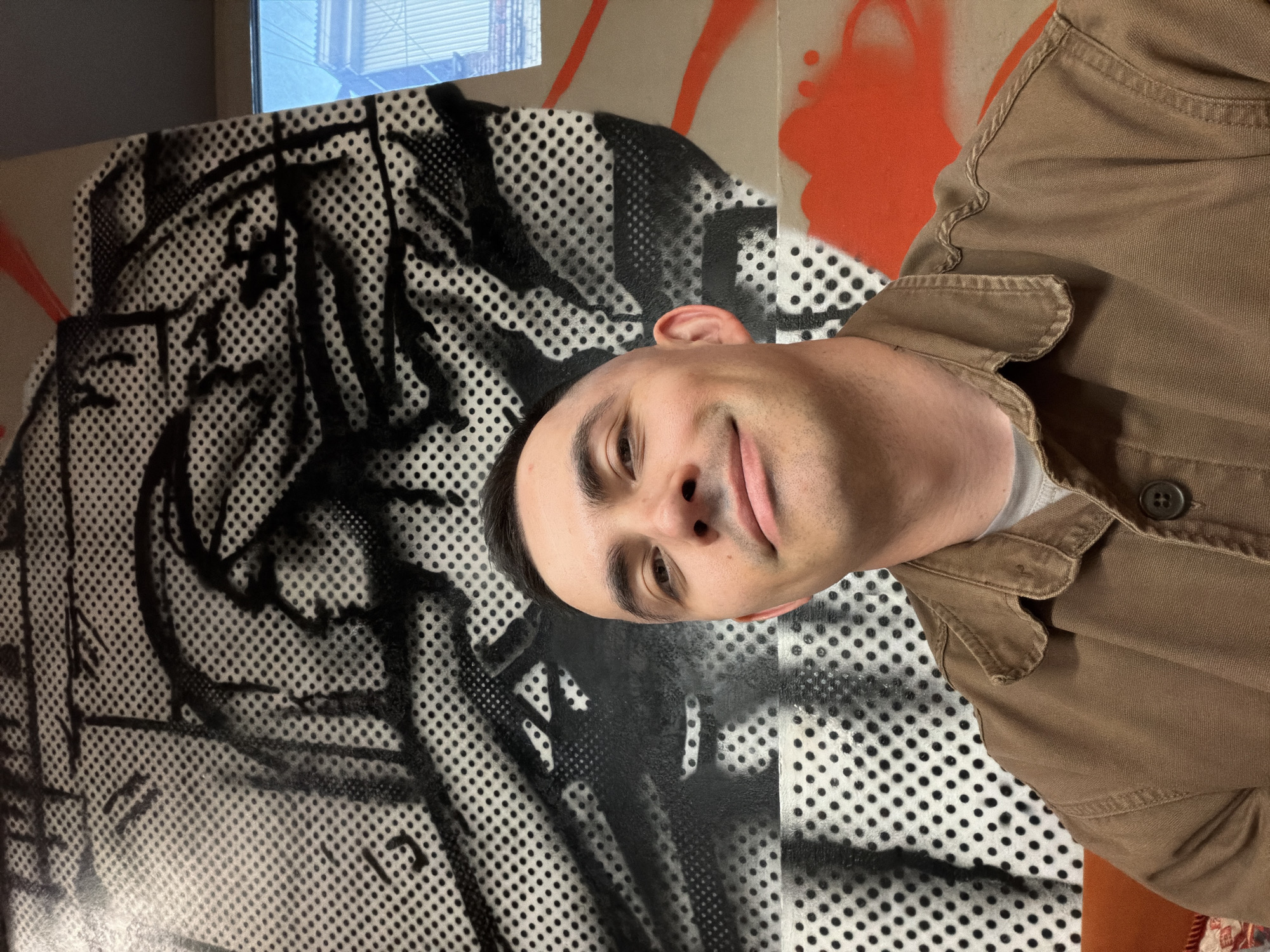 A man with short dark hair and a light complexion is posing in front of a piece of modern art that uses black and white dots. He is wearing a brown collared shirt. In the background, there is what appears to be orange graffiti. The location is not specified as Izhevsk, Russia, nor is there evidence of it in the image.