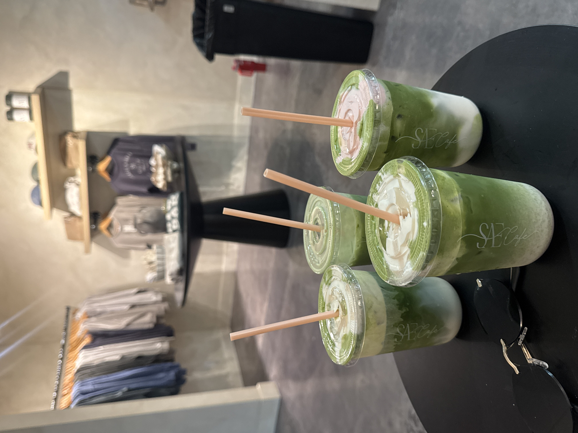 In a retail environment, likely a cafe or boutique in Hollywood, California, four green beverages with white creamy toppings and orange straws are arranged on a black circular table. The drinks are in clear plastic cups, with a subtle white logo on one of the cups that appears to read "SAE Cafe". In the foreground, a pair of black sunglasses rests on the table. In the background, a rack of clothing is visible, along with shelving units displaying merchandise and two dark-colored trash cans. The lighting suggests an indoor setting, possibly during daylight hours.