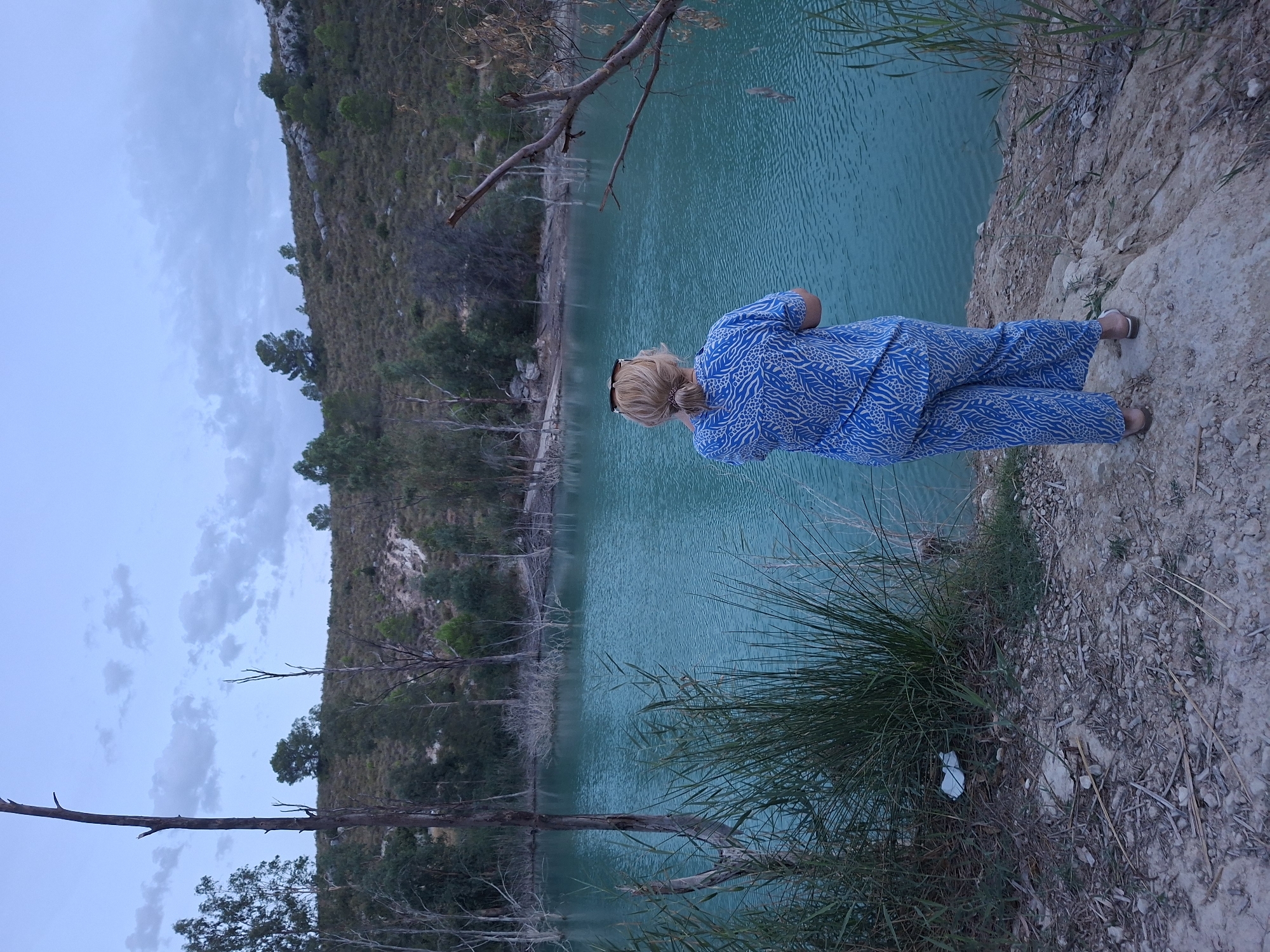 In the city of Mula, Spain, a woman stands facing a bright turquoise body of water, possibly a lake or reservoir, surrounded by a dry, hilly landscape. She is wearing a blue patterned pajama set and sandals. The water's edge is rocky and covered with dry grasses and some scattered debris, including a white plastic bag caught in the vegetation. The background features a barren, tree-dotted hillside under a cloudy twilight sky. The overall scene suggests a remote, natural setting.