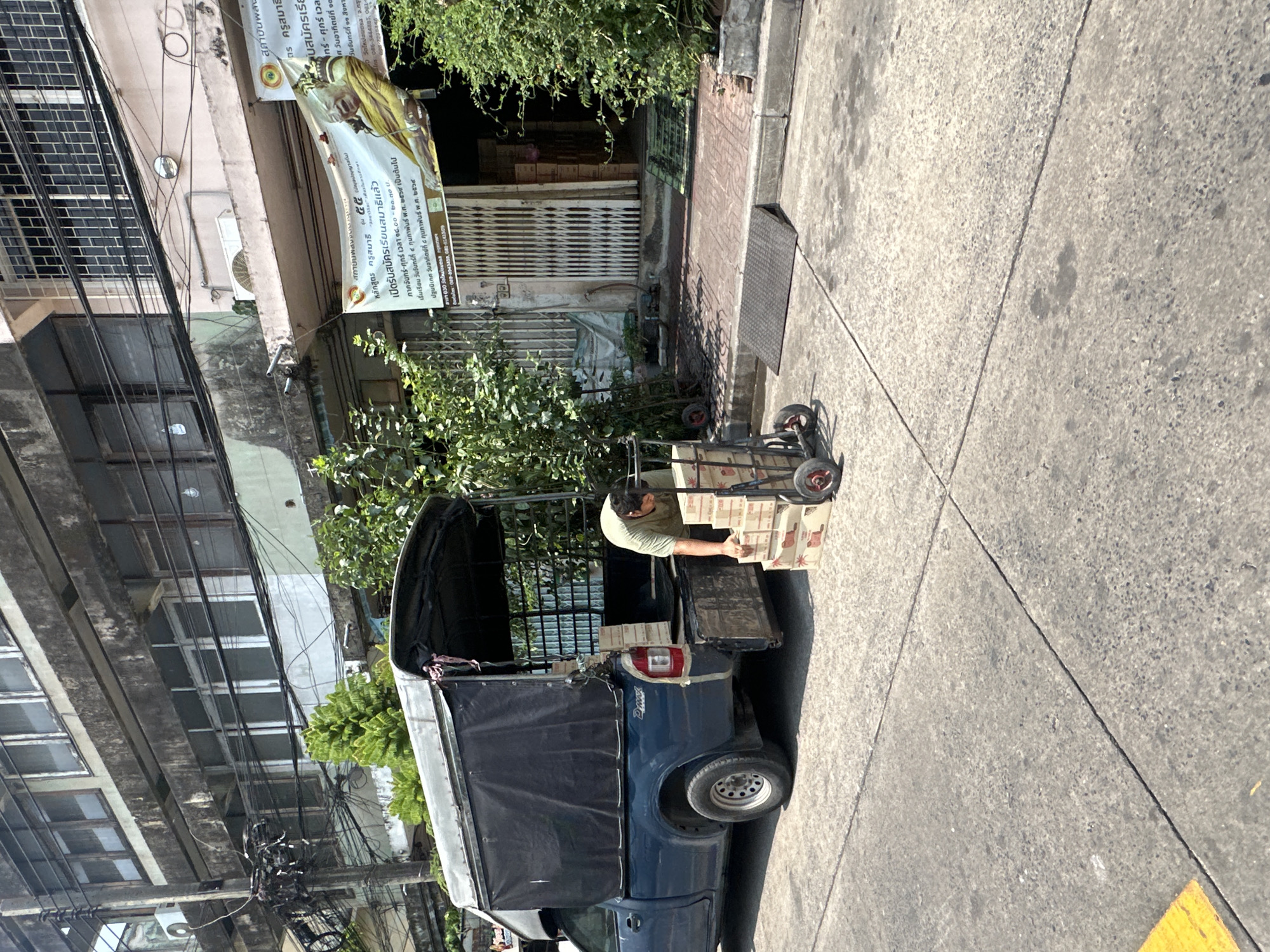 The image captures a street scene in Bangkok, Thailand. A man is unloading boxes from the back of a blue "tuk-tuk" vehicle. The vehicle has a black canvas cover. The boxes are stacked on a small wheeled cart. Behind the vehicle, there is a building with an open storefront, and adjacent to it, a sign in Thai script. The street is concrete. Above, there is a building with multiple windows and a dense network of power lines. The lighting suggests a bright, sunny day.