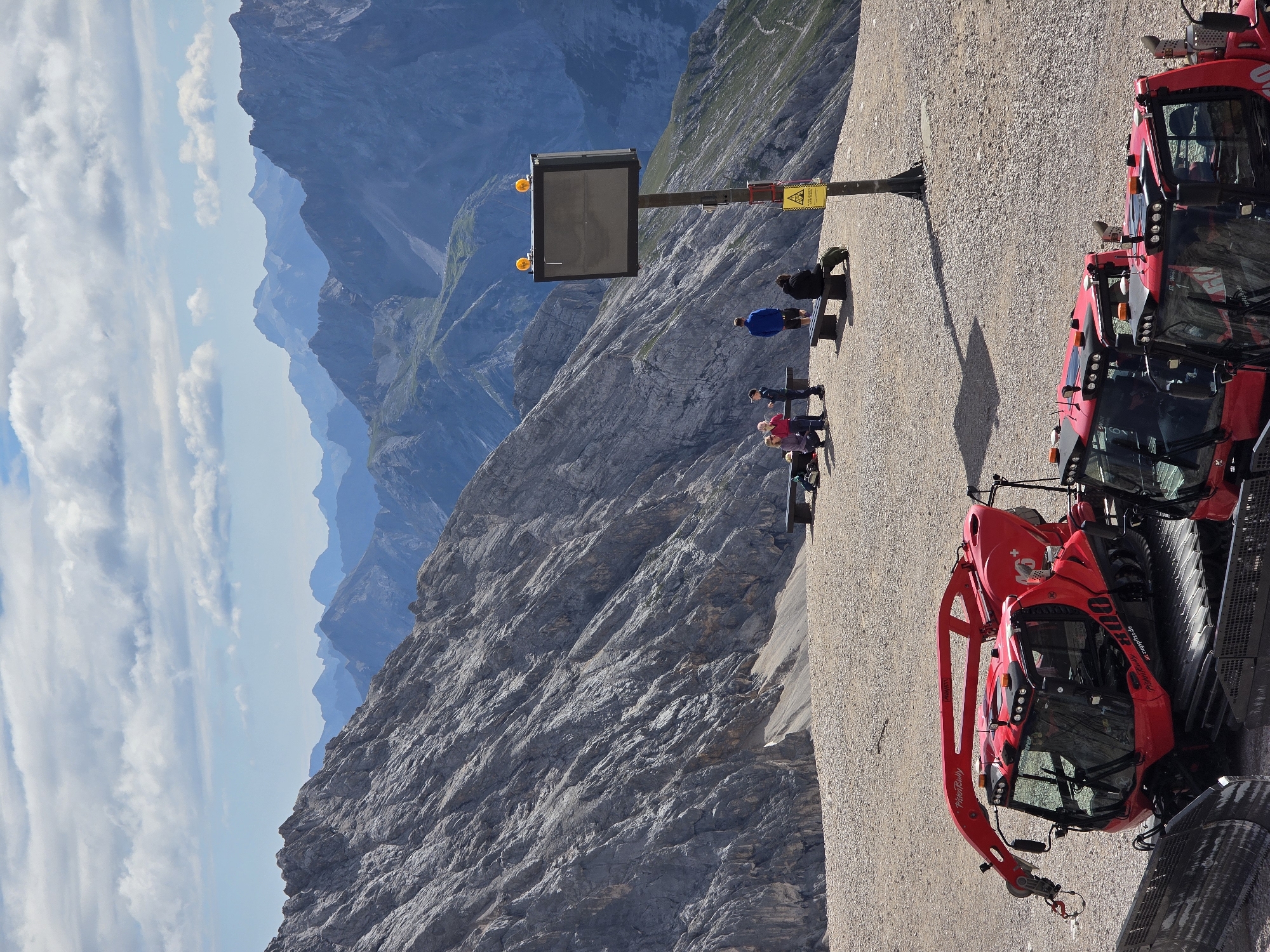 The image depicts a high-altitude mountain landscape, likely in or near Garmisch-Partenkirchen, Germany, given the presence of "Zugspitze.de" branding on a vehicle. In the foreground, two bright red snow groomer vehicles are parked on a gravelly surface. The larger one on the left features a prominent articulated arm. Further back, a group of people are visible, some seated on benches and others standing, with a large digital display screen mounted on a pole nearby. The background is dominated by rugged, rocky mountain ranges under a partly cloudy sky. The terrain suggests an alpine environment, possibly a tourist area or a ski resort at high elevation.