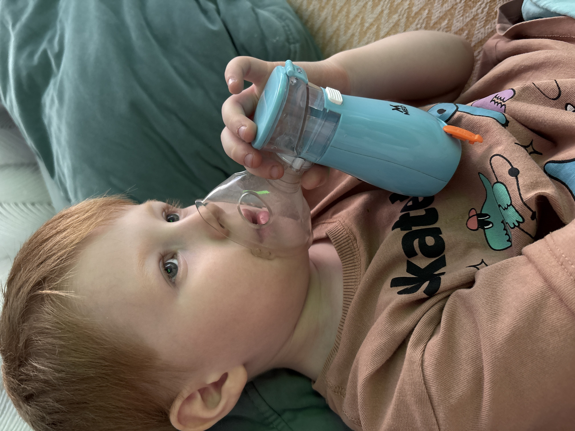 A young, fair-skinned child with red hair and blue eyes is lying down, facing the camera. The child has a clear, plastic nebulizer mask covering their nose and mouth. An adult hand holds a light blue nebulizer machine connected to the mask. The child wears a light brown long-sleeved shirt with cartoon dinosaur images and black lettering. The child rests on a green cushion, part of which is visible in the lower left corner. A light orange cushion is also partially visible near the top of the image. The photograph was taken indoors, and there is no information to suggest this photograph was taken in Izhevsk, Russia.