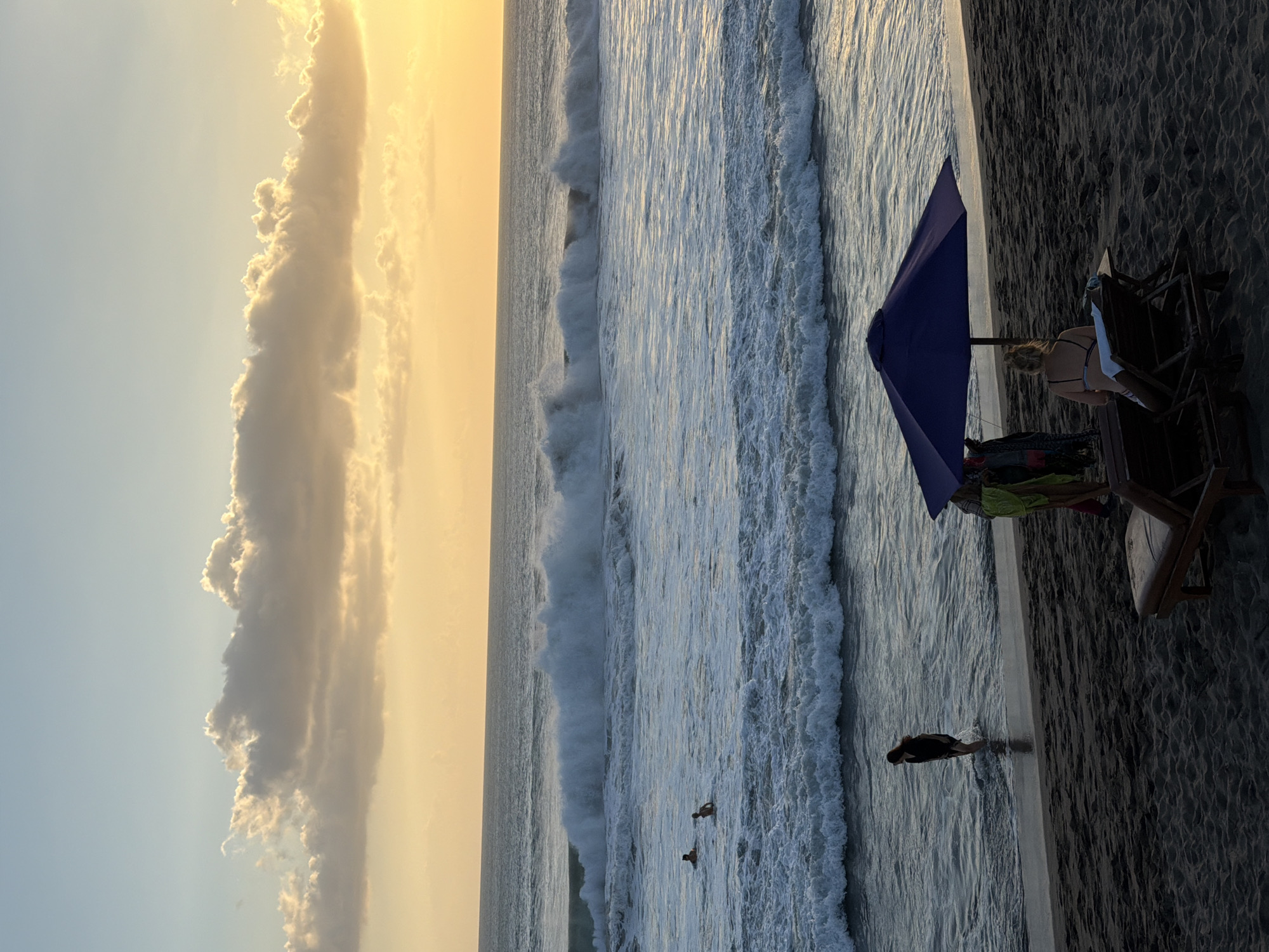 The image depicts a beach scene at sunset in North Kuta, Indonesia. The sky is a gradient of soft yellow and pale blue, with large, textured clouds illuminated by the setting sun. The ocean is active with significant waves breaking and crashing towards the shore.
In the water, several people are visible; two individuals are closer to the shore, wading or swimming, while two others are further out, appearing to be swimming or playing in the waves.
On the dark sand beach, a blue umbrella provides shade. Beneath it, a group of people, possibly vendors, are gathered, wearing colorful clothing, including a bright green garment. To the right of the umbrella, two lounge chairs are set up. One is empty, while the other is occupied by a person wearing a bikini, facing the ocean. Closer to the water's edge, a solitary figure, a woman in dark clothing, walks into the surf. The sand appears dark and textured, indicating it might be volcanic sand common in some Indonesian beaches. The overall atmosphere is tranquil, with the powerful ocean contrasting with the calm of the beachgoers enjoying the end of the day.