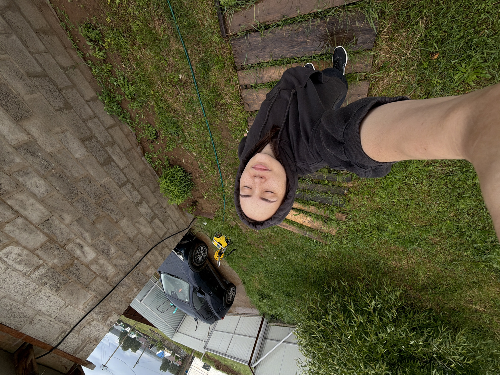 The image is an overhead selfie, rotated on its side, featuring a young adult with closed eyes and freckles, wearing a dark hooded sweatshirt. Her arm is extended upwards, holding the camera. She stands on a pathway of wet wooden planks laid over damp grass, with her feet, clad in black sneakers, visible.
To the left of the person, a diagonal gray brick wall with a textured surface defines one edge of the area. A black hose or cable runs along the base of this wall. Below the person, a dark-colored car, possibly a hatchback, is parked on what appears to be a paved or dirt surface, and it also shows signs of being wet. Next to the car, a yellow and black pressure washer or similar outdoor cleaning device is visible, suggesting recent car cleaning activity.
The ground is a mix of grass, dirt, and some weeds, all appearing damp, indicating recent rain. A large, leafy green bush occupies the lower right corner of the frame. In the background, behind the car, there's a metal fence or gate, and distant buildings, power lines, and poles are visible under an overcast sky. The scene depicts an outdoor, somewhat rural or suburban backyard setting in Chaikovsky, Russia, during daytime under cloudy weather conditions. No discernible text is visible.