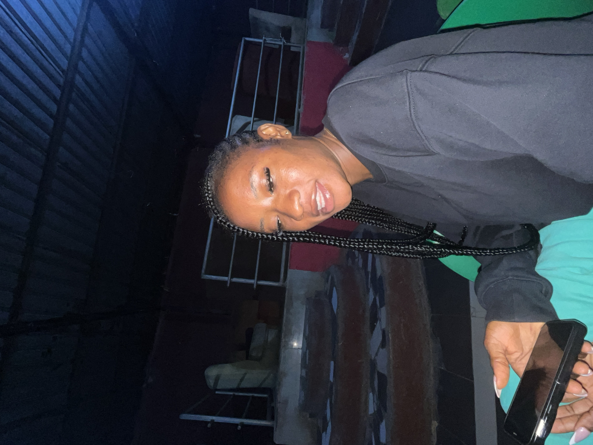 An outdoor night scene depicts a young Black woman sitting and holding a smartphone. She has long, braided dark hair and is wearing a black sweatshirt and turquoise pants. The background features a dark, corrugated metal roof, a red wall, and tiered concrete steps with decorative tiling. A metal railing is visible behind the woman. The location is specified as Gura Top, Nigeria.