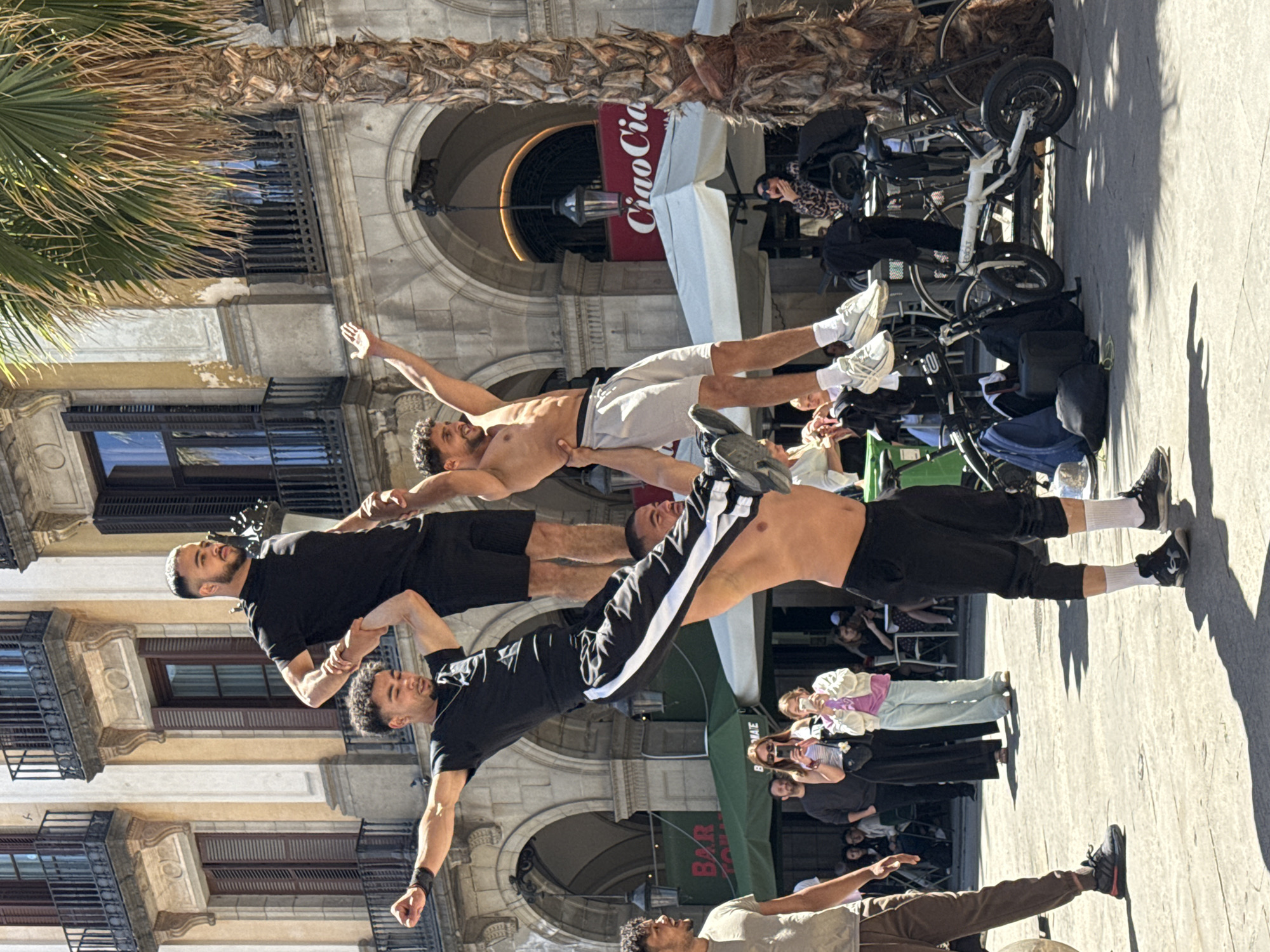 A performance art tableau vivant features four men arranged in a vertical stack in an urban setting. The men are posed with the intention of creating a living sculpture. The bottom man is standing. The next three men are positioned horizontally above the first man. The second man is in black short sleeves and shorts. The third man is shirtless, wearing white shorts. The top man is wearing a white t-shirt and tan pants. People are watching the performance. Bicycles are visible in the background. Architectural details of the building include arched walkways and balconies. A sign for “Ciao Ciao” is also in the background. This performance art takes place in Barcelona, Spain.