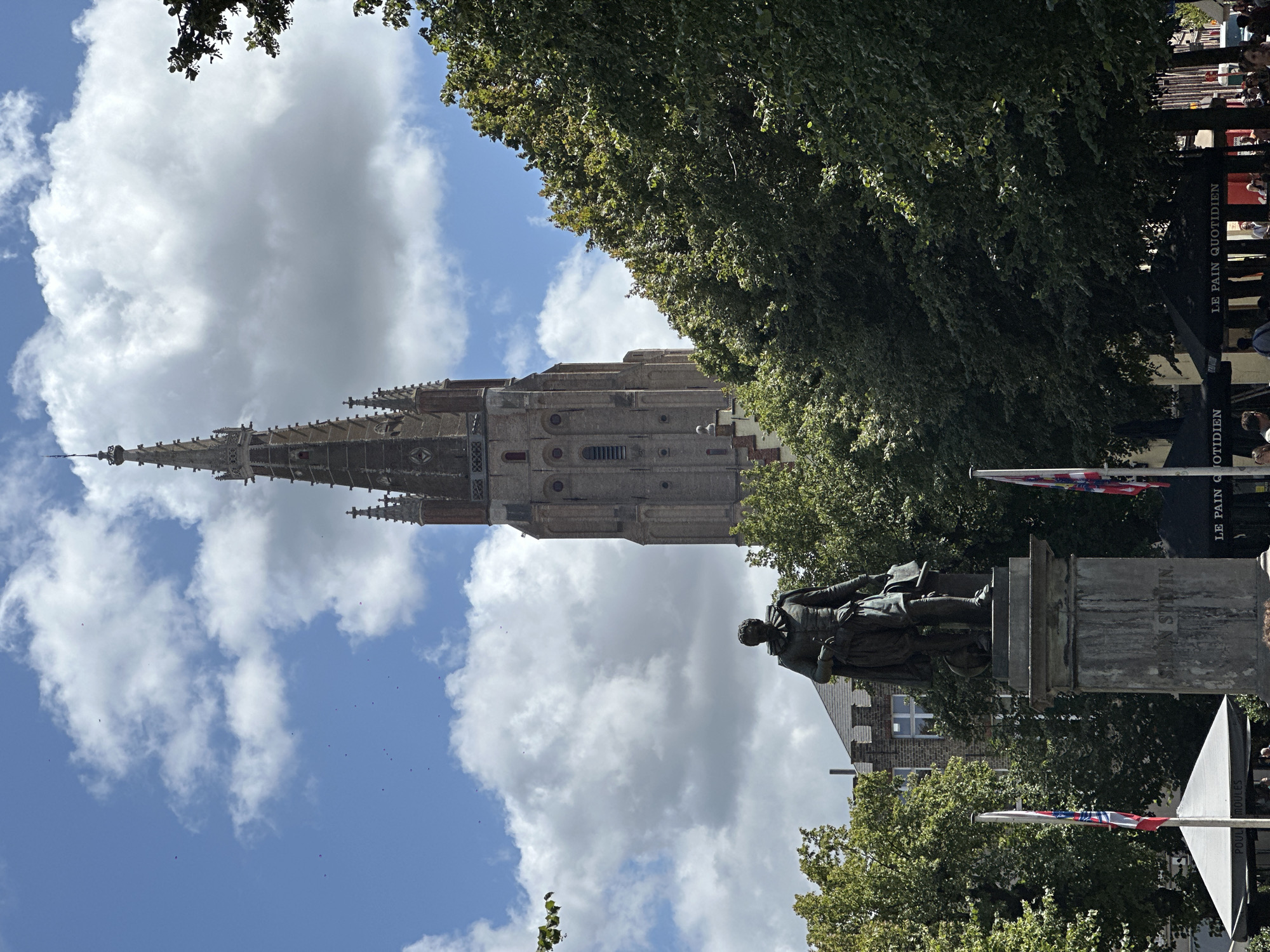 The image depicts a sunny day in Bruges, Belgium, with a towering brick church spire dominating the background. In the foreground, a bronze statue of a man stands on a stone pedestal, likely a historical figure from the city. The statue is surrounded by lush green trees, and flags of various countries are visible, suggesting a public square or monument area. Cafes with outdoor seating are also present, indicating a lively atmosphere. The sky is a vibrant blue, with scattered white clouds adding depth and texture to the scene.