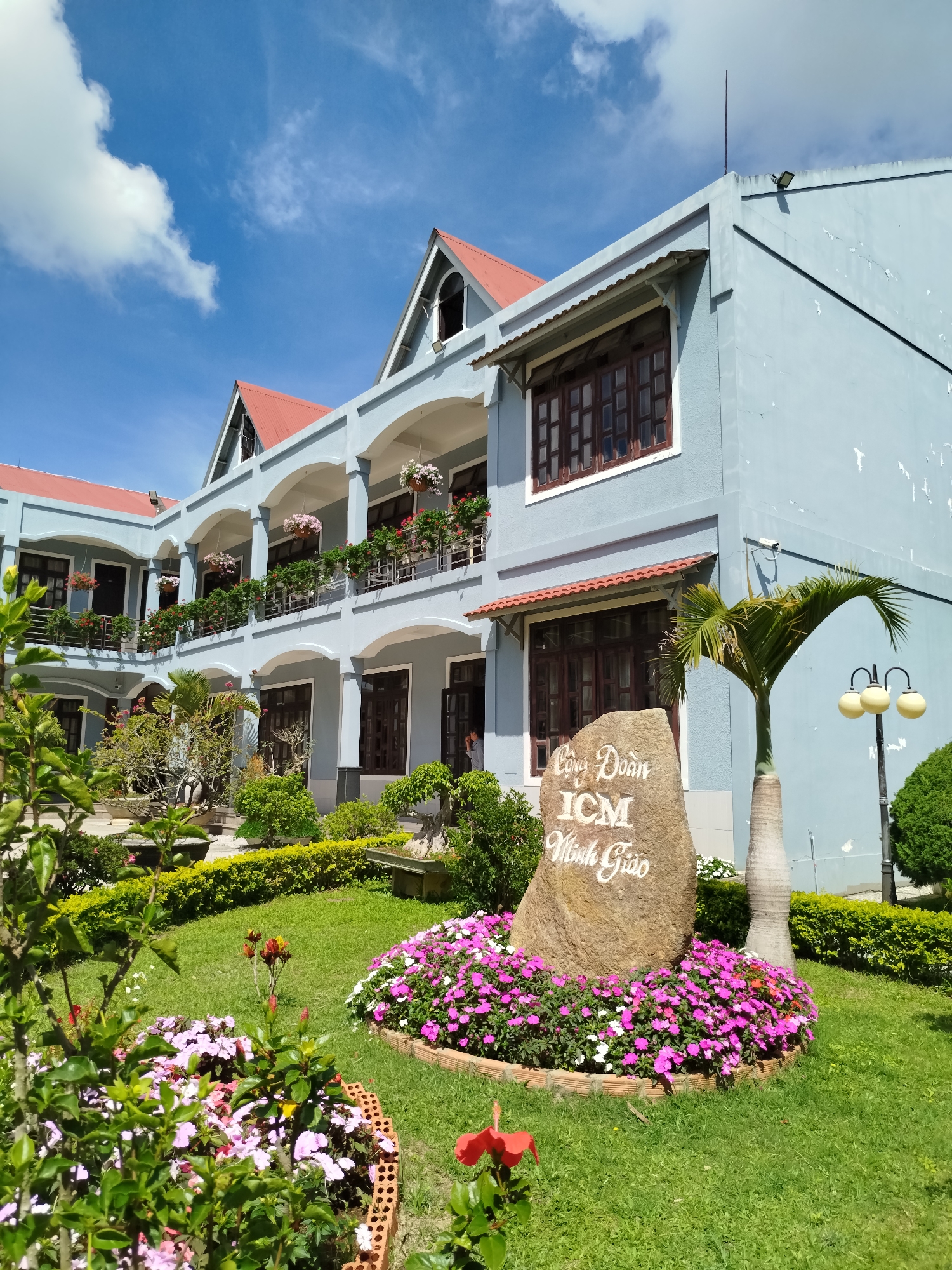 The image shows a two-story building in Da Lat, Vietnam, with a light blue facade, red roofing, and arched balconies adorned with flower boxes. A stone monument stands in front of the building, bearing the inscription "Cong Doan ICM Minh Giao." A palm tree and a vintage lamppost are situated to the right of the monument. The foreground features a well-maintained garden with pink and white flowers, trimmed hedges, and various plants. The sky is clear with scattered clouds. The building appears to be in a well-kept, serene setting.