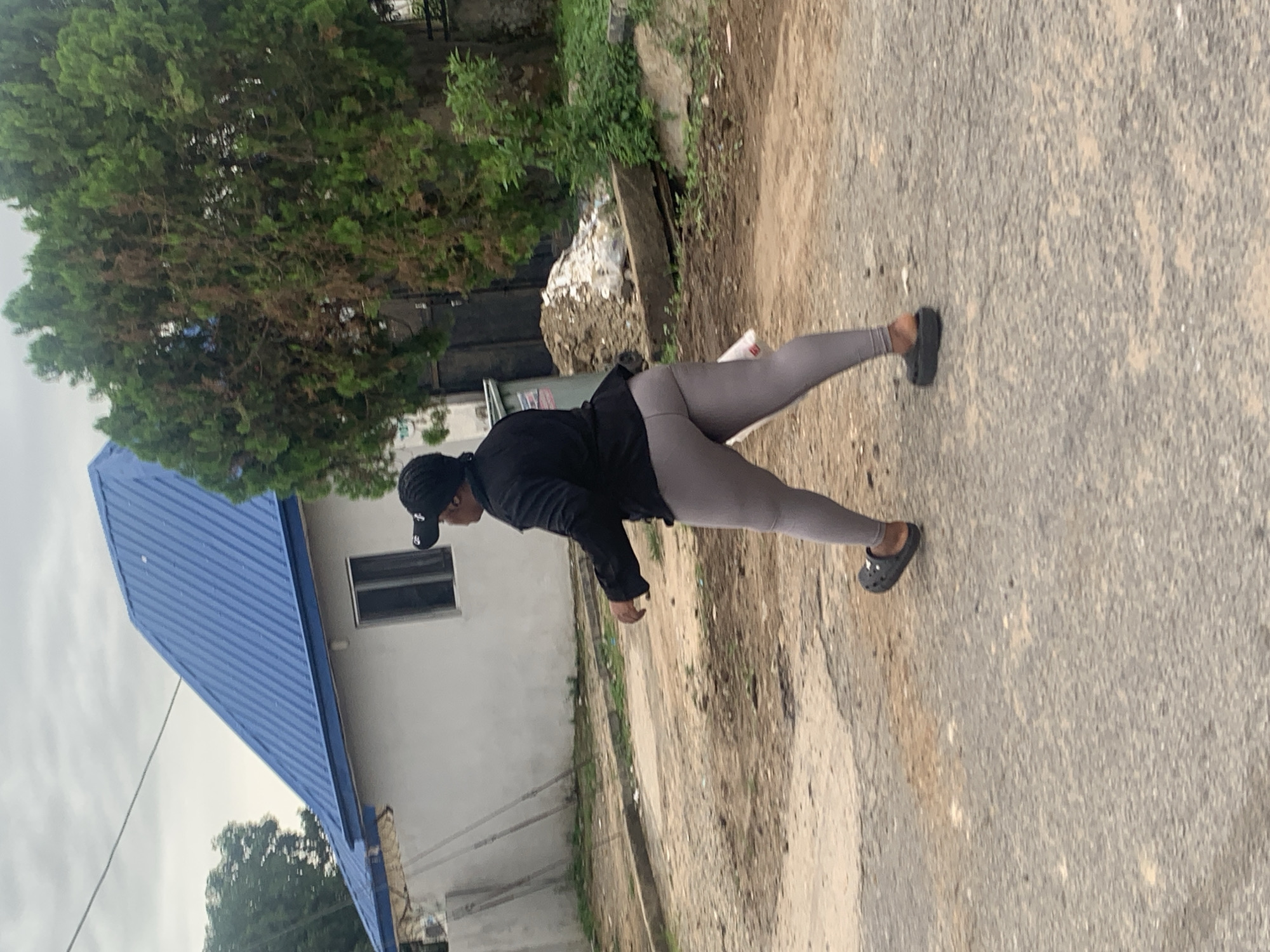 A woman wearing a black jacket, grey leggings, and black crocs walks away from the camera on an overcast day. She is wearing a black baseball cap. In the background, a white building with a blue corrugated metal roof is visible. To the right of the building, a dense green tree stands. The ground is a mix of dirt and asphalt. The scene appears to be in a residential area of Abuja, Nigeria.