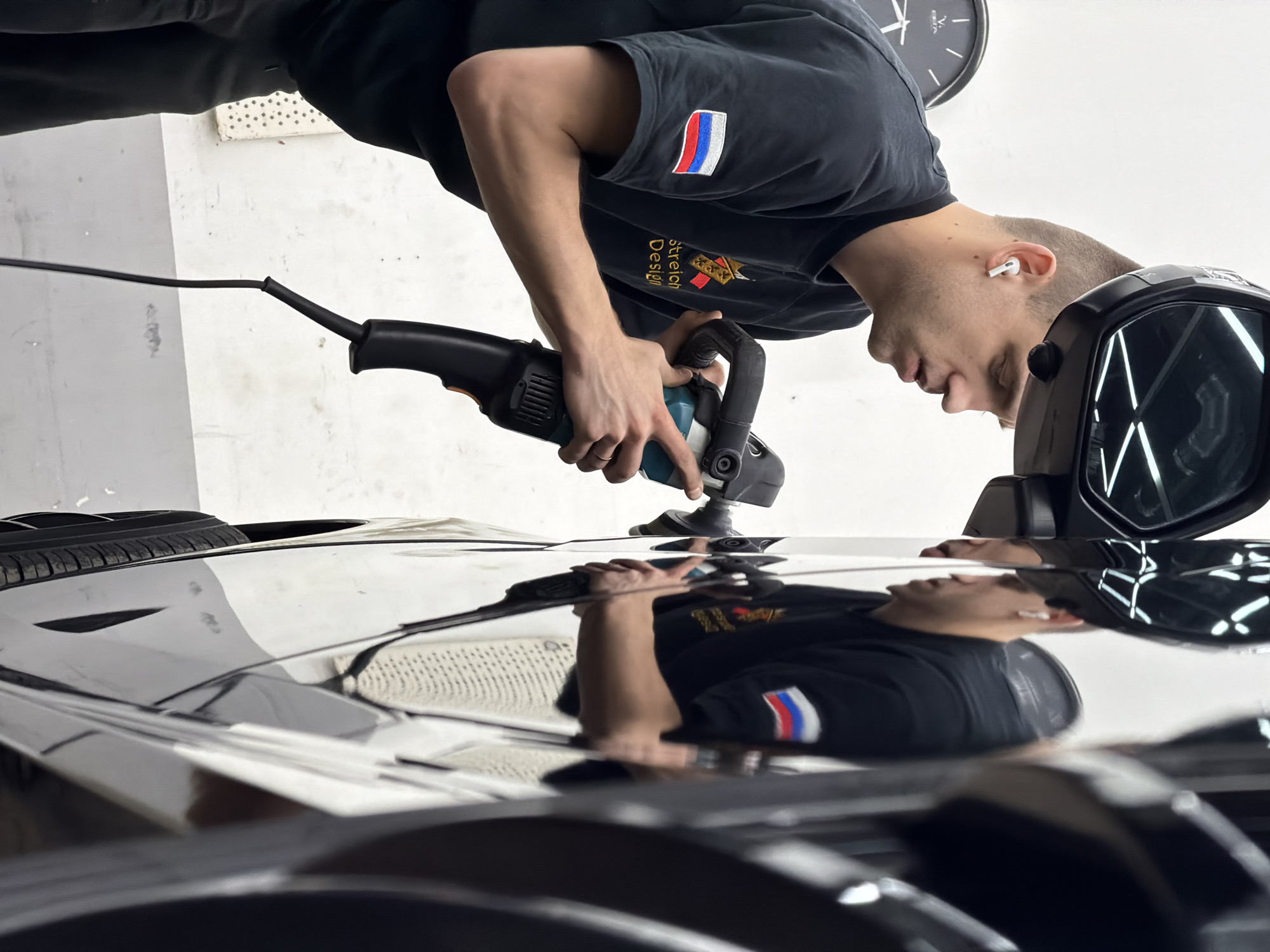 A man is polishing the hood of a black car. The man is wearing a black t-shirt with a Russian flag patch on the sleeve and wireless earbuds in his ear. He is using a handheld polisher. The hood of the car reflects the man and the surrounding environment. The location appears to be indoors, possibly a garage or auto detailing shop. A clock is visible on the wall in the background. Given the Russian flag patch, the location is likely in Russia, but I lack specific information to confirm the city as Krasnoyarsk.