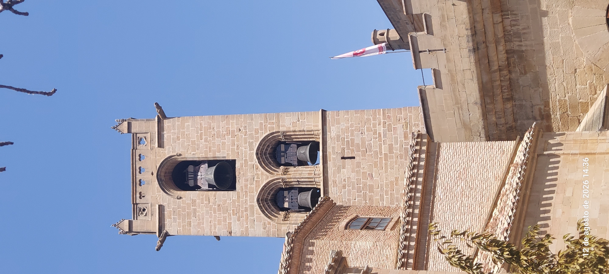 The image shows the bell tower of the Palace of the Kings of Navarre in Olite, Spain, under a clear blue sky. The tower is constructed of stone and features Gothic architectural elements, including arched openings for the bells, gargoyles, and decorative battlements. Two large bells are visible in the upper archway, and two smaller bells are in the archway below. To the left of the bell tower is a brick building with a tiled roof. To the right, a stone wall with a flag flying from a pole can be seen. The flag is white with a red cross and some red insignia. The lower part of the image shows a tree branch with leaves and a timestamp overlay that reads "12 de agosto de 2026 14:36". The scene is one of architectural grandeur and historical significance, bathed in bright sunlight.