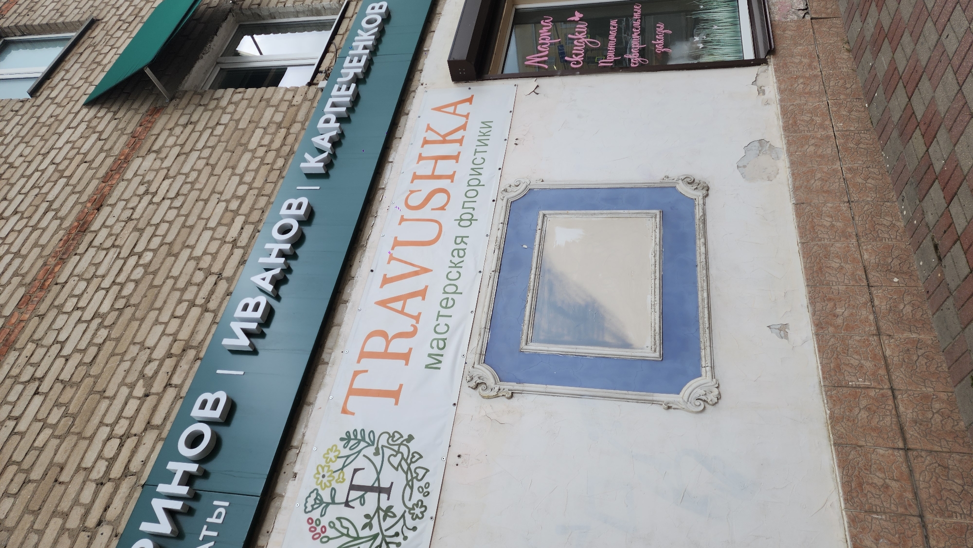 The image shows the exterior of a building in Smolensk, Russia. Several signs are attached to the building: a green sign with white Cyrillic lettering reading "ГРИНОВ | ИВАНОВ | КАРПЕЧЕНКОВ"; a banner reading "TRAVUSHKA" (in orange) and "мастерская флористики" (in green), which translates to "floristry workshop" in English, alongside a floral design; and a sign in a window mentioning "марта скидки" (March discounts) in pink Cyrillic letters. A blue decorative frame is mounted on the wall. The building's facade appears aged, with peeling paint and wear. The photograph seems to have been taken from a low angle. Part of a brick wall and a tiled area are visible at the edges of the frame.