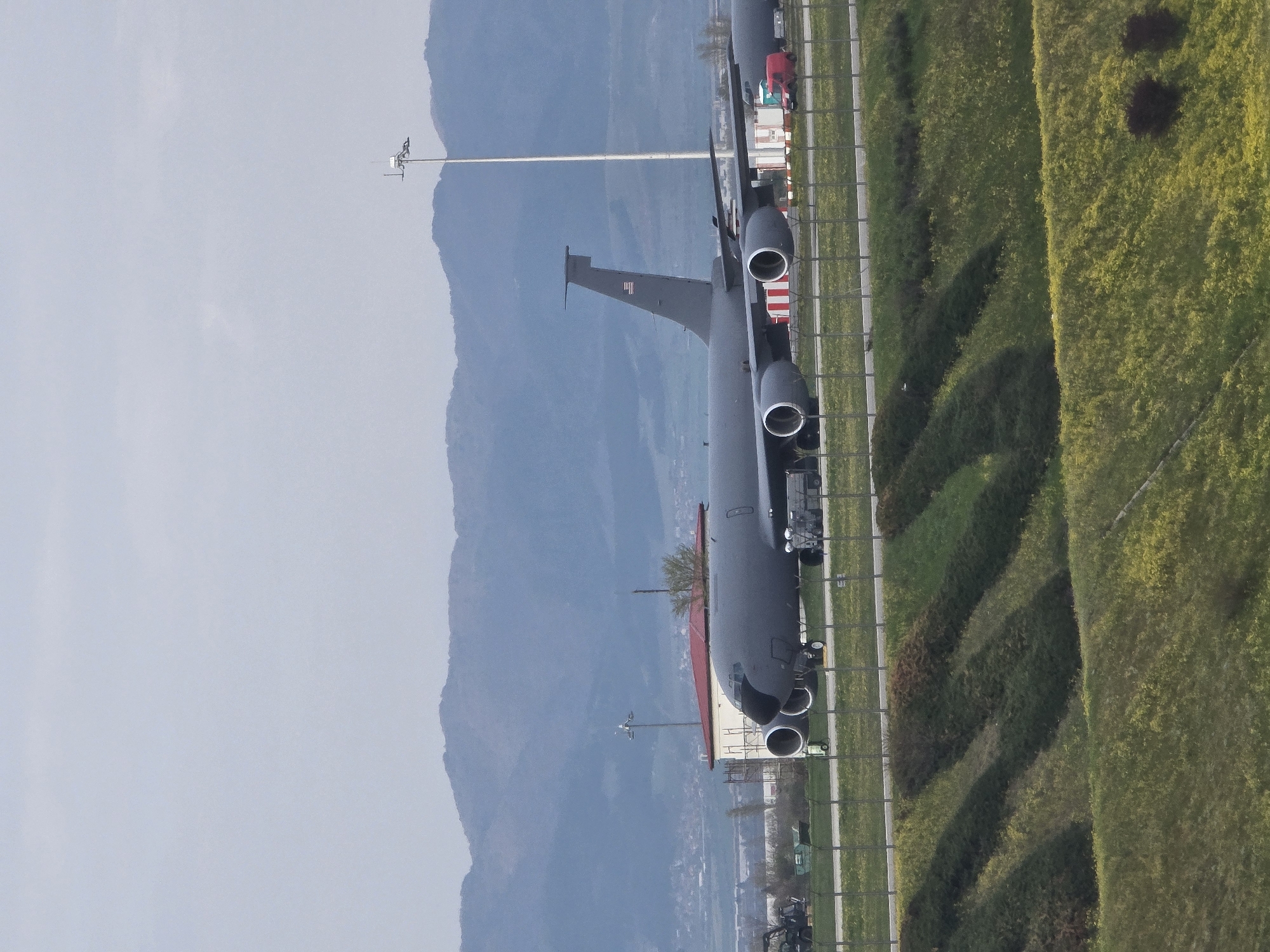 The image shows a large grey military cargo plane parked on an airfield adjacent to a green, grassy area with foliage. In the background, there is a mountainous landscape. The setting might be near the city of Sofia, Bulgaria, given the terrain. Details of other infrastructure, such as a possible road and a small building or structure, are visible parallel to the aircraft. A streetlight is positioned further back in the field.