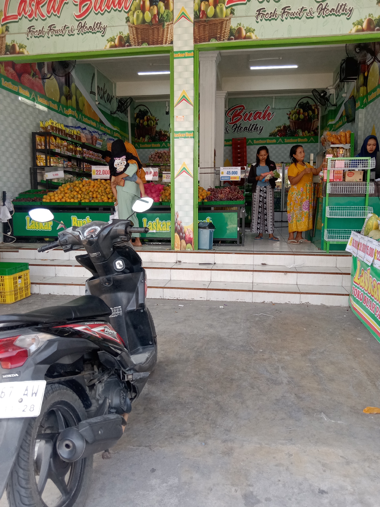 The image depicts a fruit stand in Klepek, Indonesia. A black Honda motorcycle is parked in the foreground. Behind it, the fruit stand displays a variety of produce, including oranges and grapes. Three women are visible at the stand, two of whom appear to be customers and one is likely an employee. The stand has signage indicating "Laskar Buah" which translates to "Fruit Army". One sign also says "Fresh Fruit & Healthy". The building appears to be a shopfront with an awning and tiled stairs leading up to the entrance. The overall environment suggests a typical street scene in a local Indonesian market.