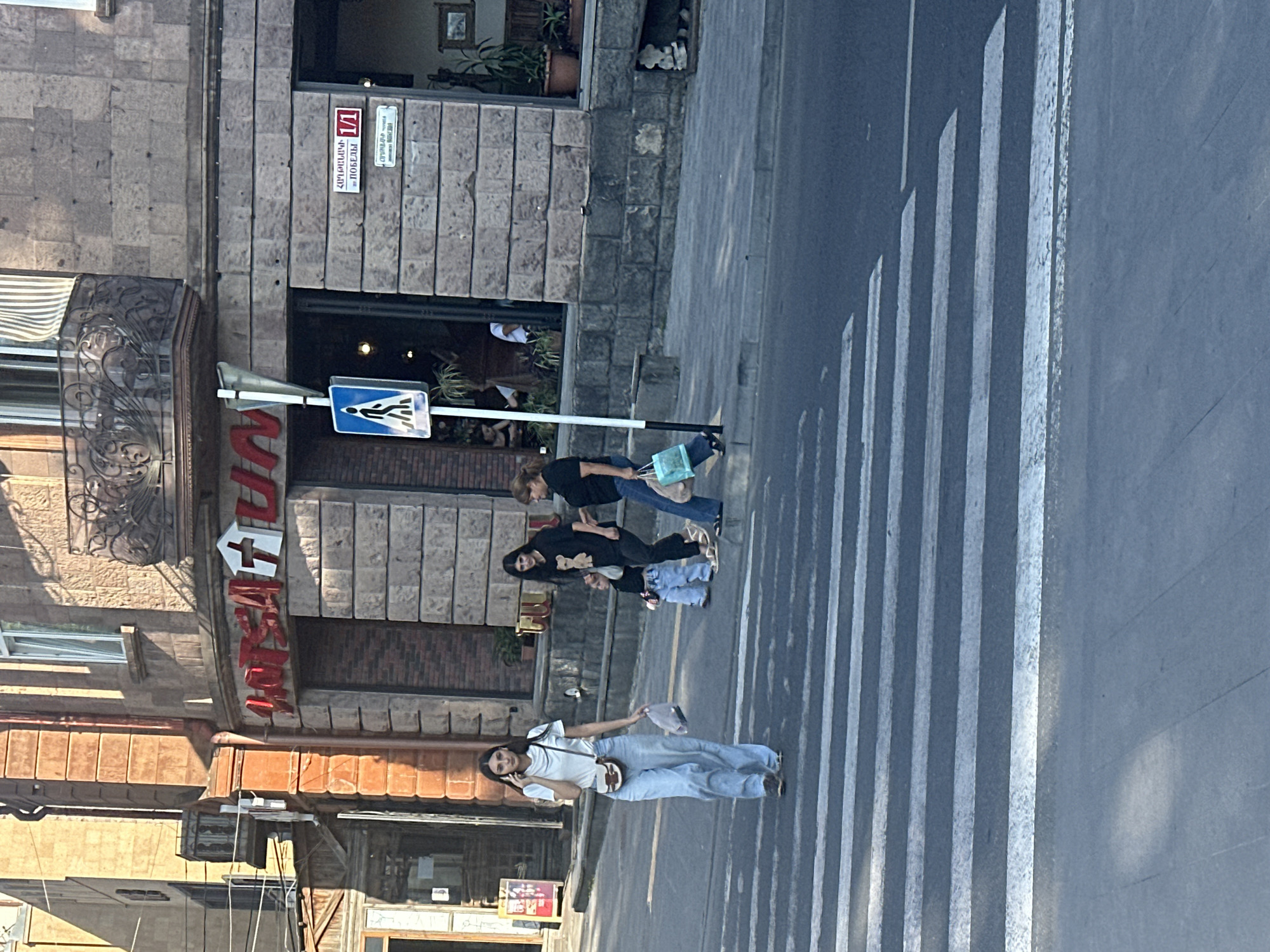 A sunny daytime street scene in Gyumri, Armenia, shows a group of people crossing a pedestrian street. The building facade is made of stone, with a balcony and a sign that says "HOTEL TUN" in red letters. A blue pedestrian crossing sign is visible. A young woman in a white shirt and light blue jeans is talking on her phone while crossing the street. Behind her, a woman in a black shirt and jeans walks with a child, and another woman in a black shirt and jeans carries a blue shopping bag. The street is paved and has white pedestrian crossing lines.