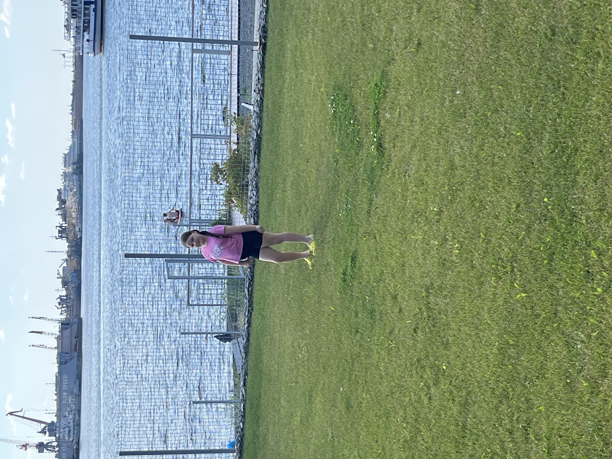 The image shows a young girl standing on a grassy area in front of a body of water. The girl is wearing a pink t-shirt with the word "MAGIC" printed on it and black shorts. She has her hair in a braid. In the background, there is a fence and beyond that, the water is choppy. Further in the background, there are several large ships and industrial structures, likely at a port. The sky is partly cloudy. The scene appears to be a waterfront recreational area. The location is identified as the embankment in the city of Chaikovsky, Russia, near the Kama River.